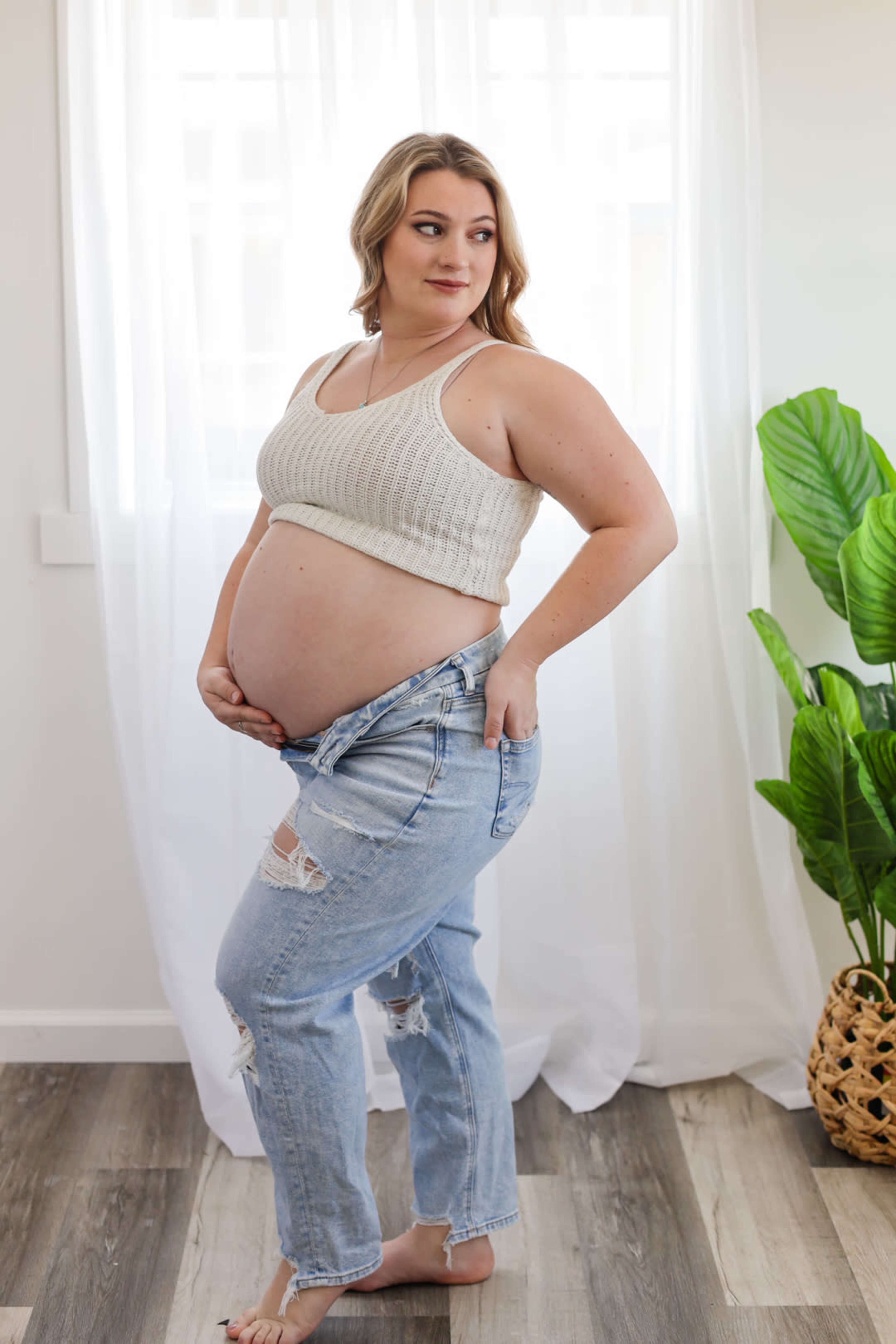 A pregnant woman stands indoors, holding her unbuttoned jeans at her hips, with a plant in the background.
