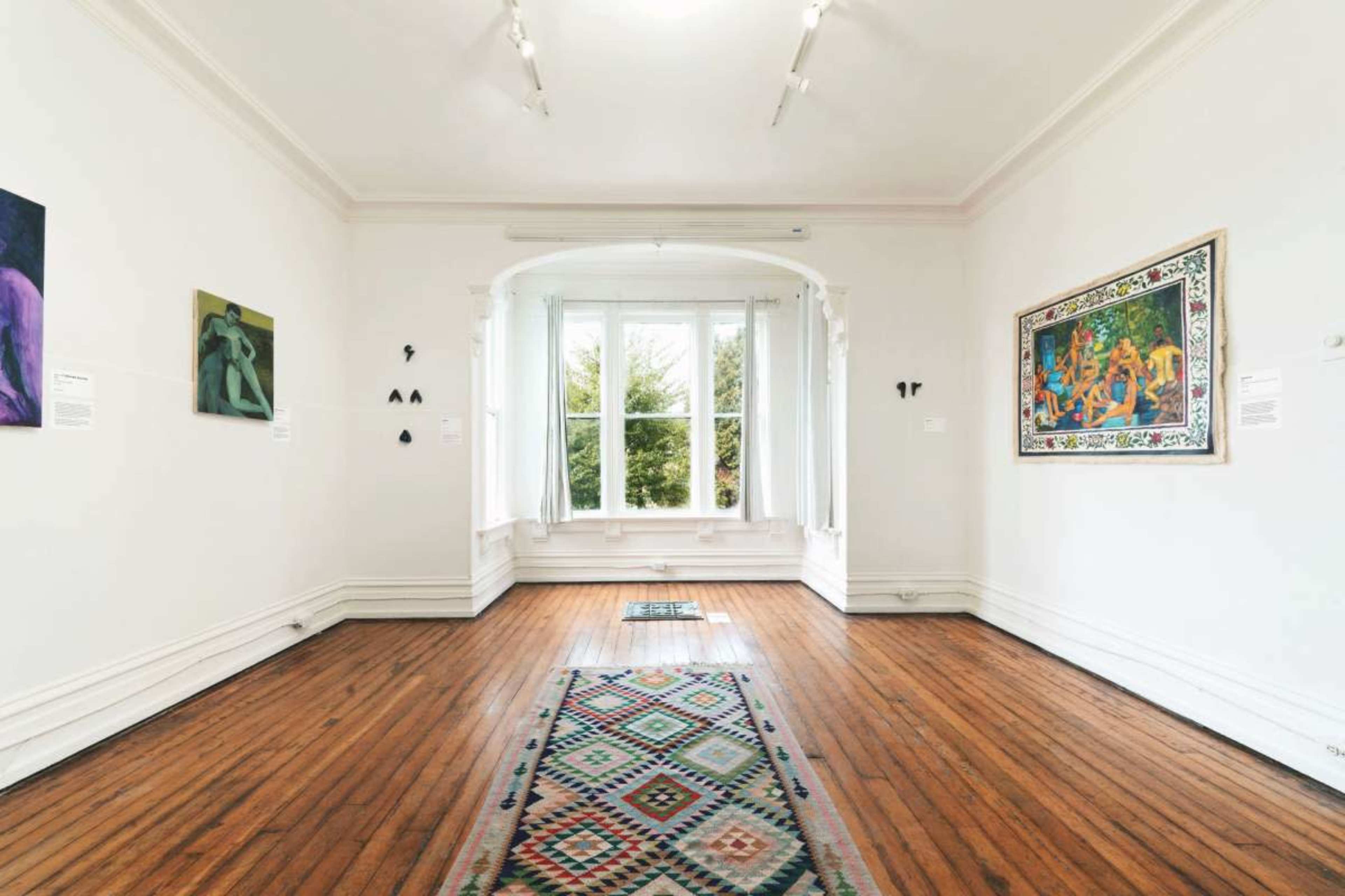 An art gallery with wooden floors, white walls, and several pieces of artwork displayed, along with a window that lets in natural light.