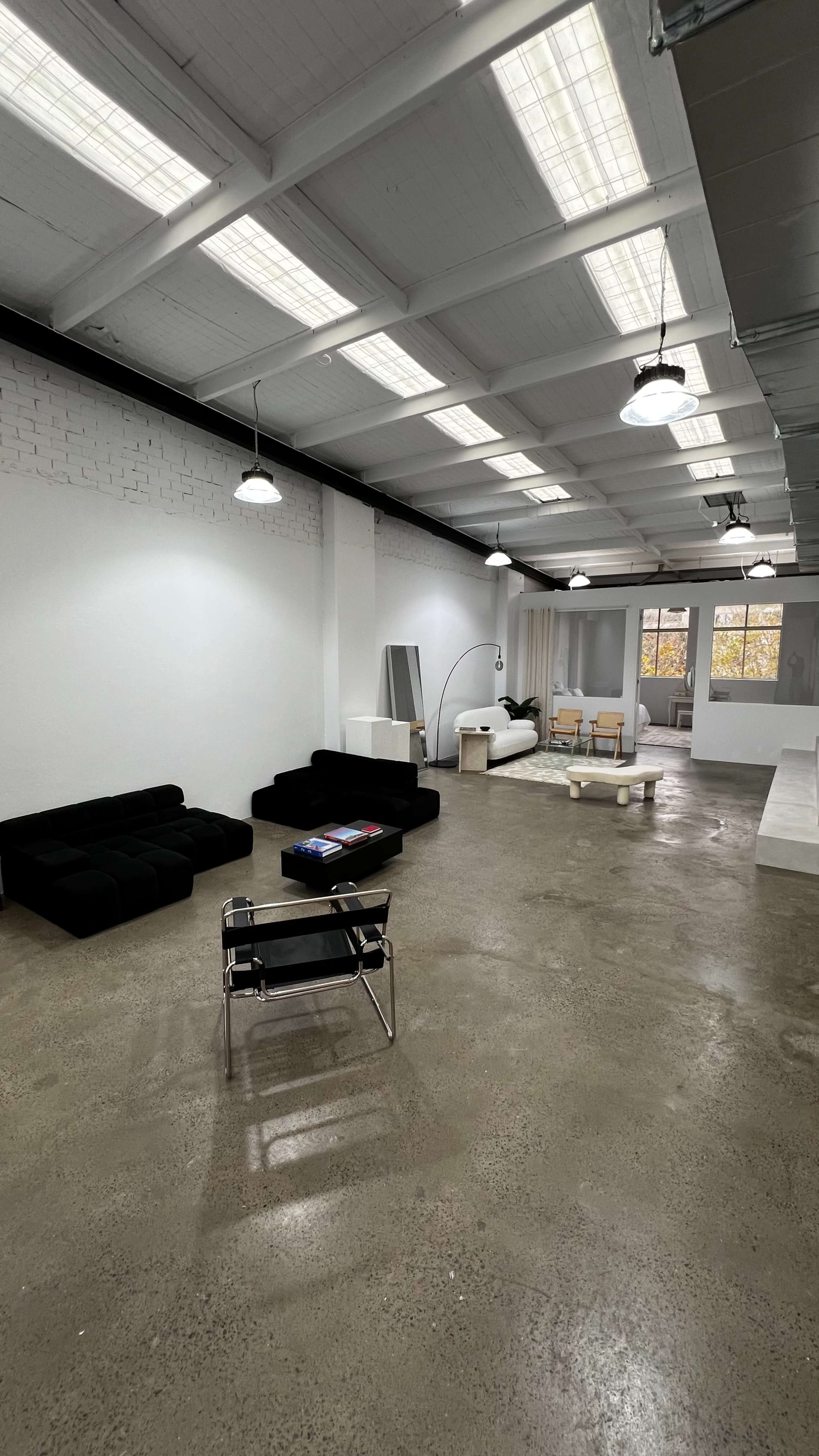 A spacious, minimalist room with two black sofas, a light wooden table, and large windows allowing natural light to enter.
