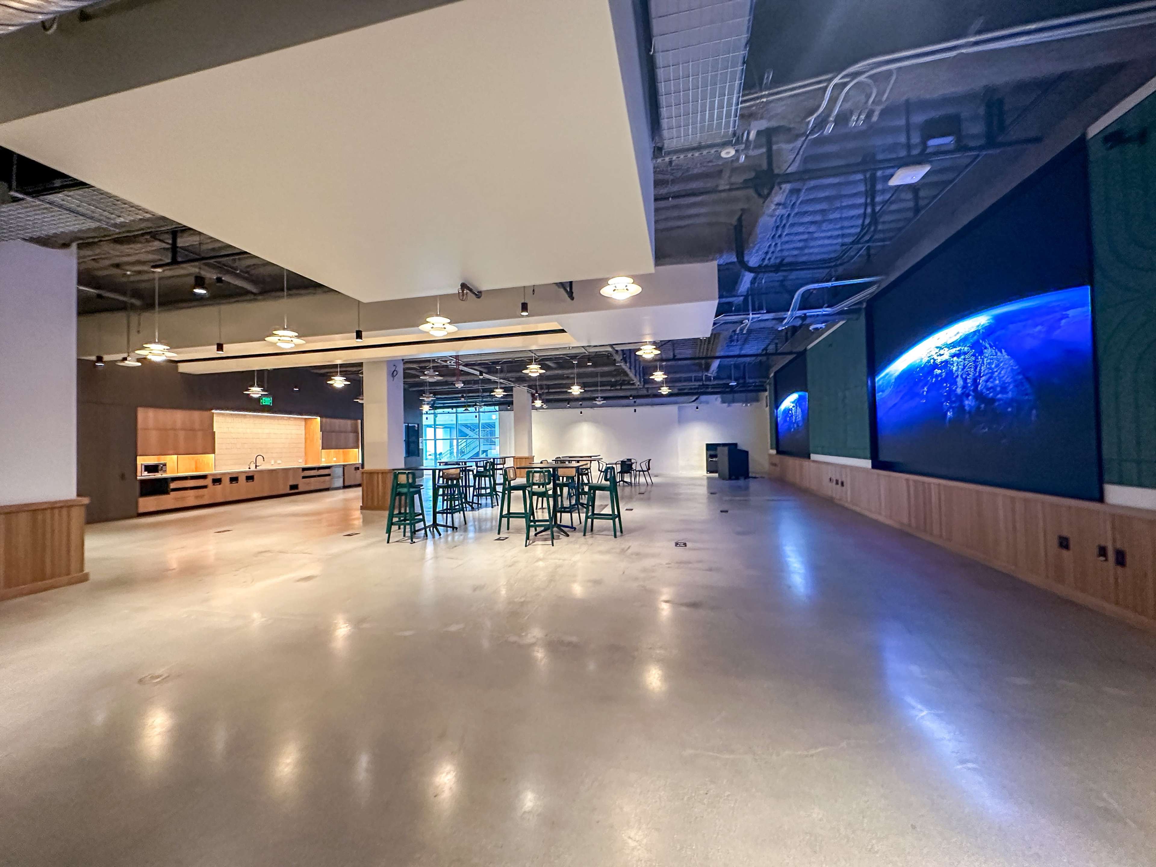 A spacious, modern interior with concrete flooring, a large screen displaying a globe, and tables and chairs arranged for gatherings.