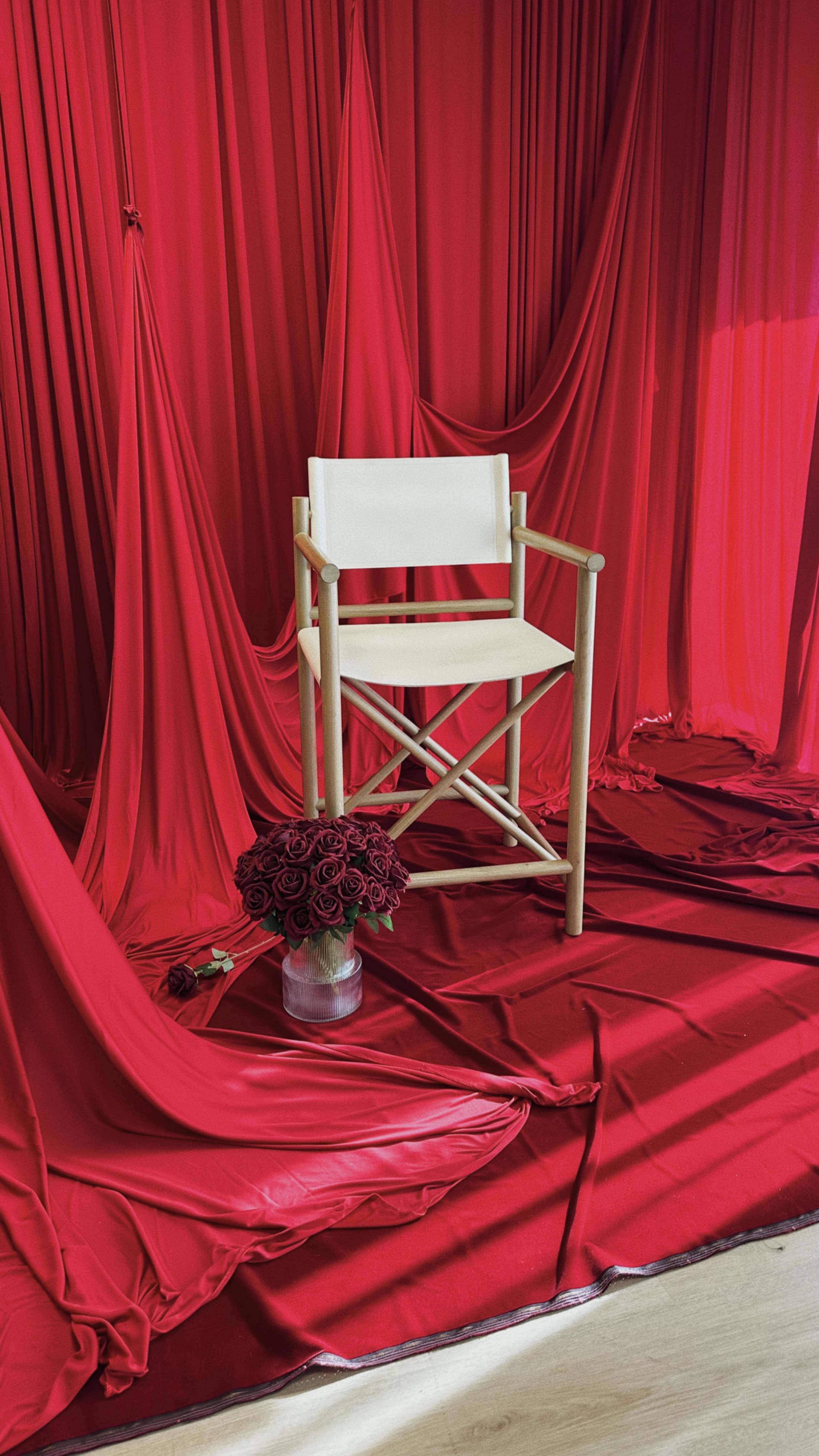 A white director's chair sits in front of cascading red drapery, accompanied by a bouquet of red roses in a glass vase on the floor.