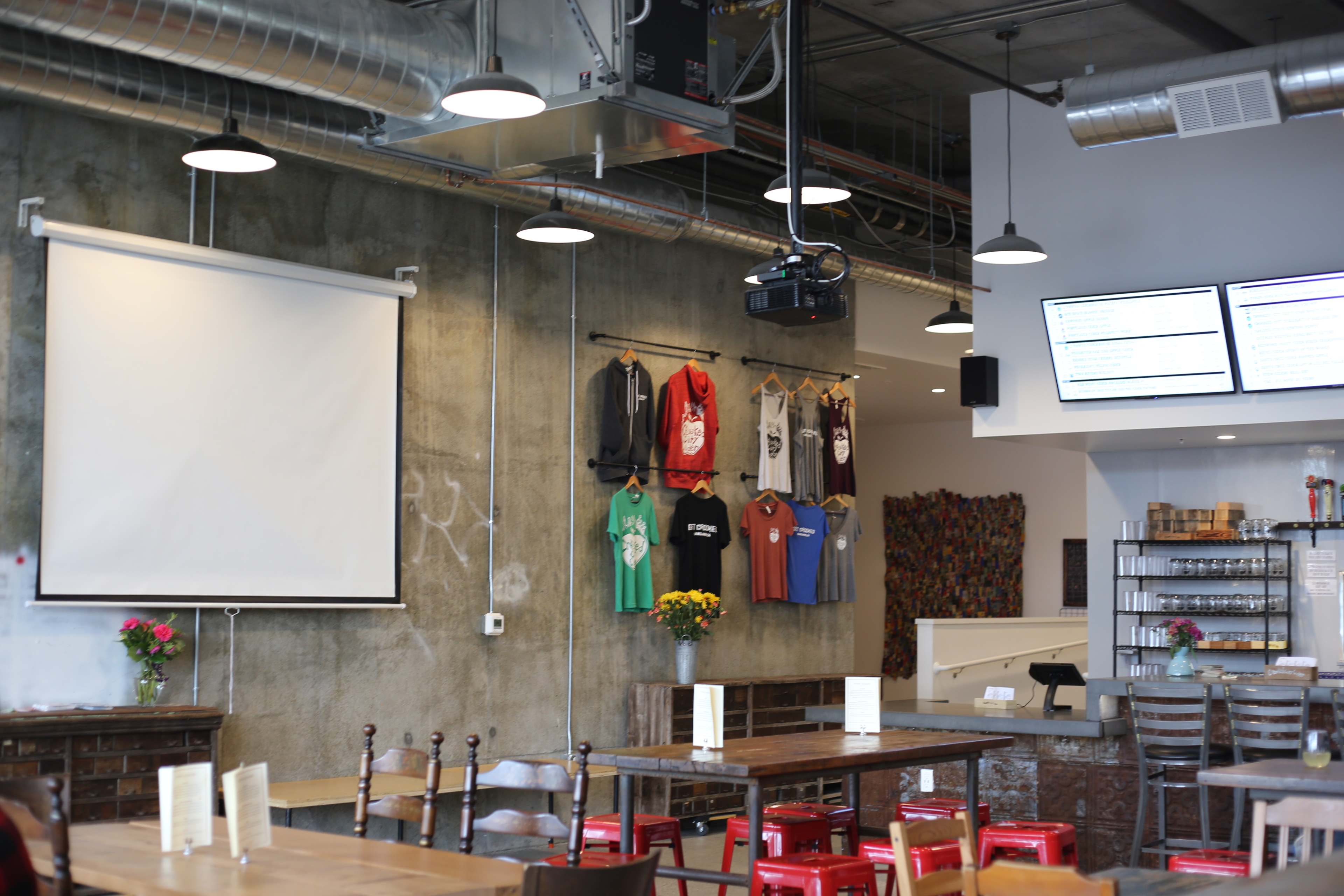 The image shows a modern café interior with various T-shirts displayed on the wall, tables with menus, and a projector screen hanging in front.