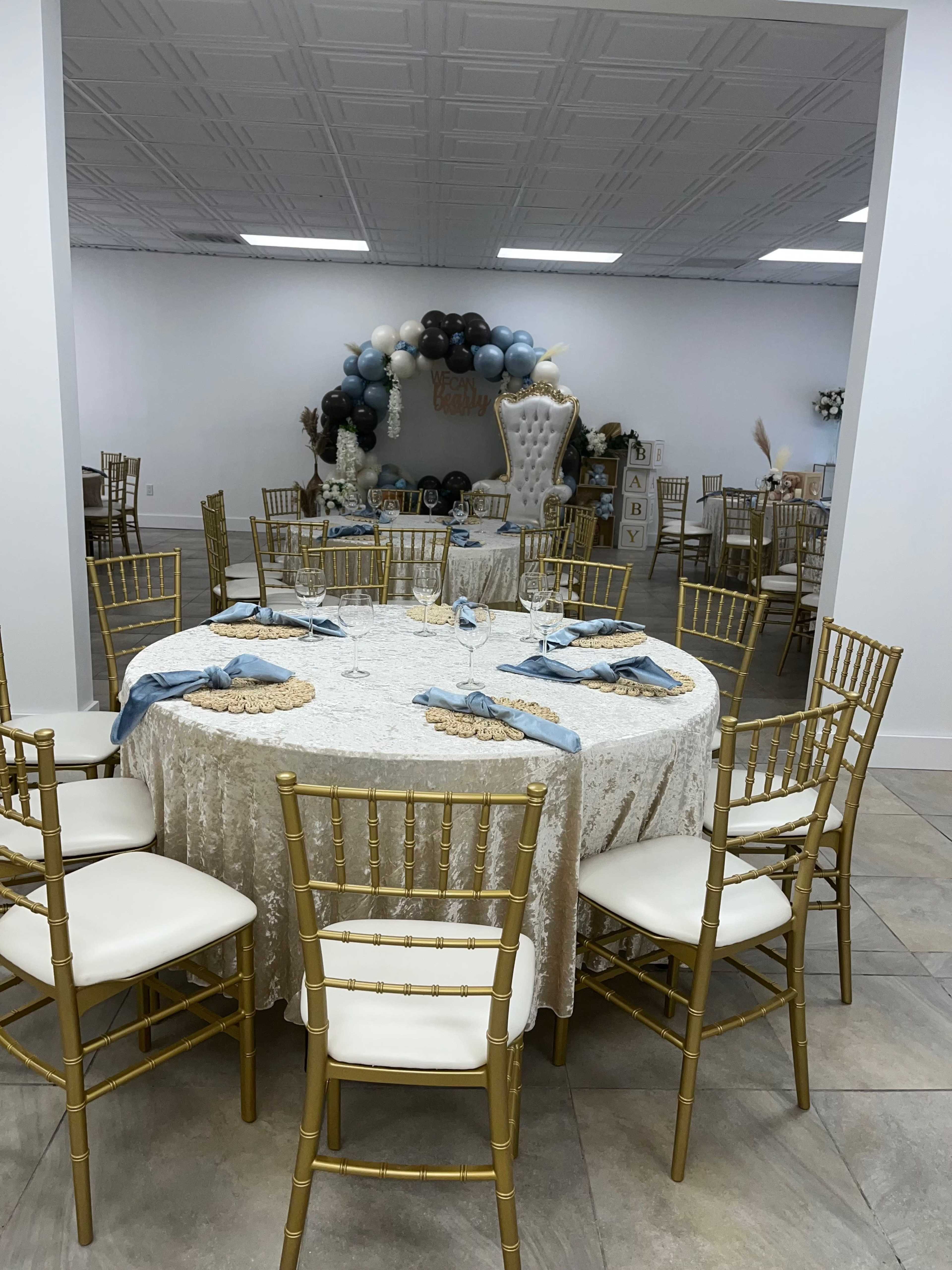 A banquet hall features elegantly set tables with gold chairs and a decorative balloon arch in the background.