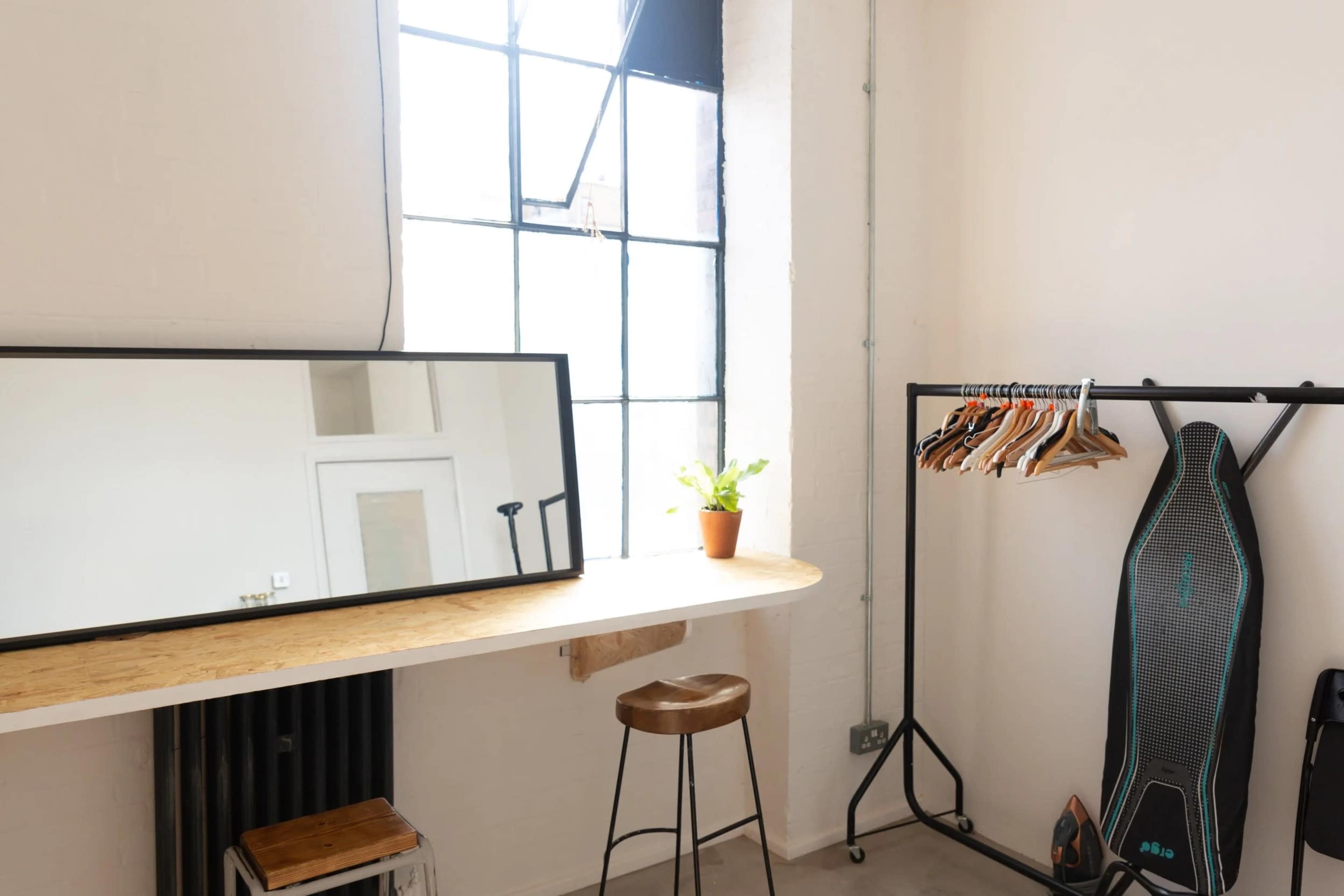 The image shows a minimalist room featuring a large mirror, a wooden countertop with a stool, a potted plant, and a clothing rack with several hangers.
