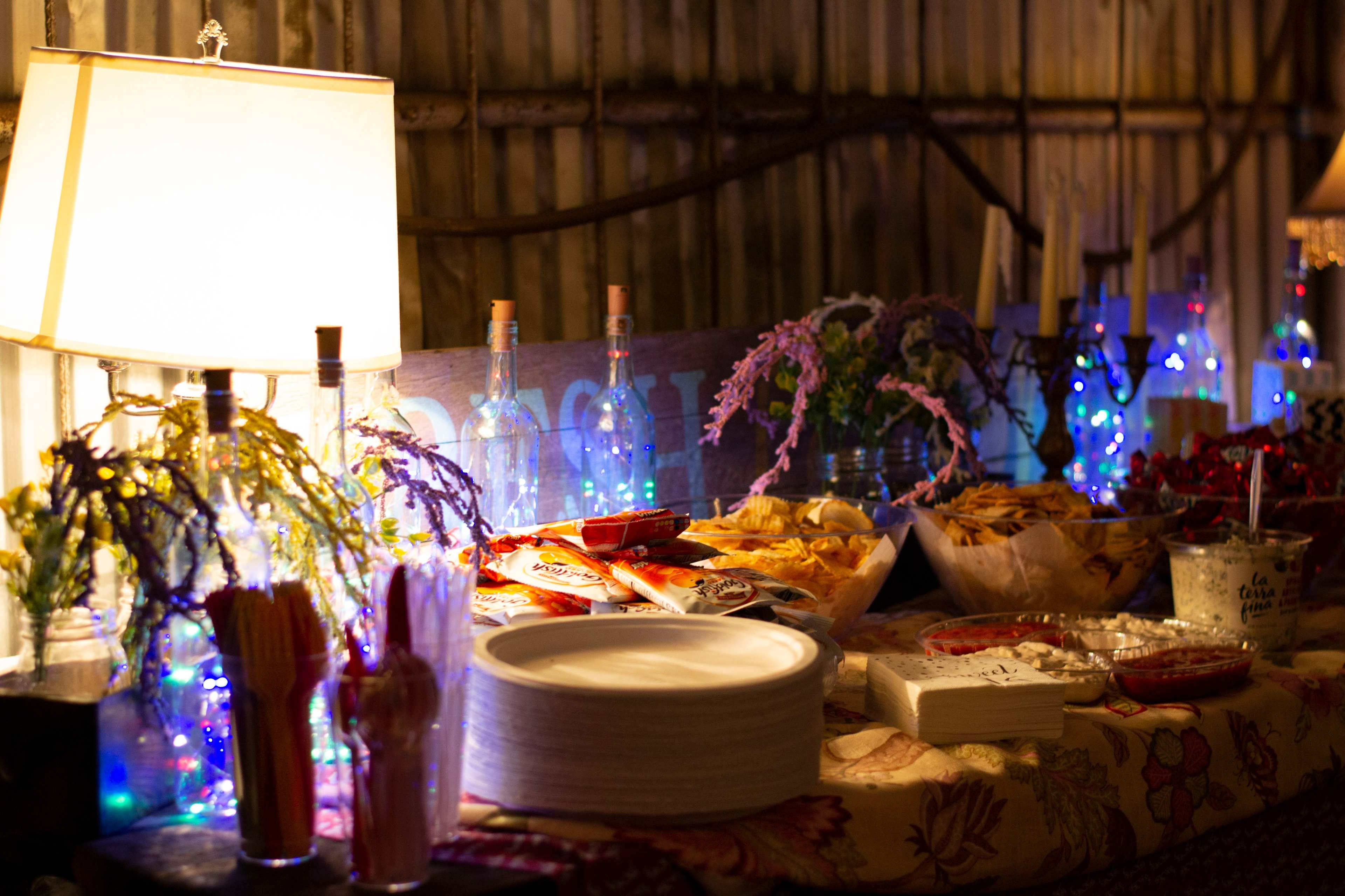 The image shows a table set up with various snacks and drinks, illuminated by lamps and decorated with colorful lights and flowers.