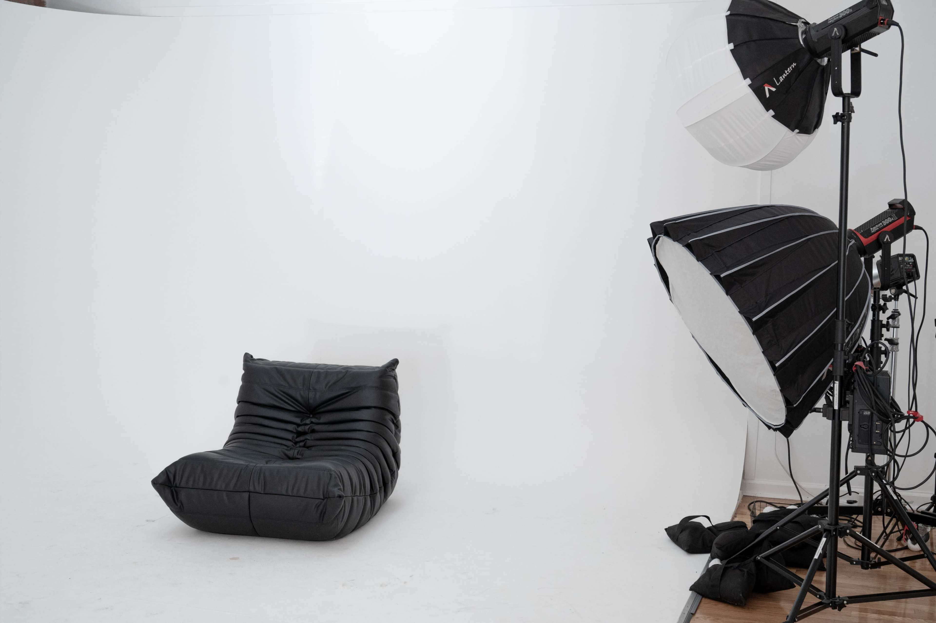 The image shows a black, modern lounge chair positioned to one side of a photography studio with a white backdrop and studio lighting equipment on the other side.
