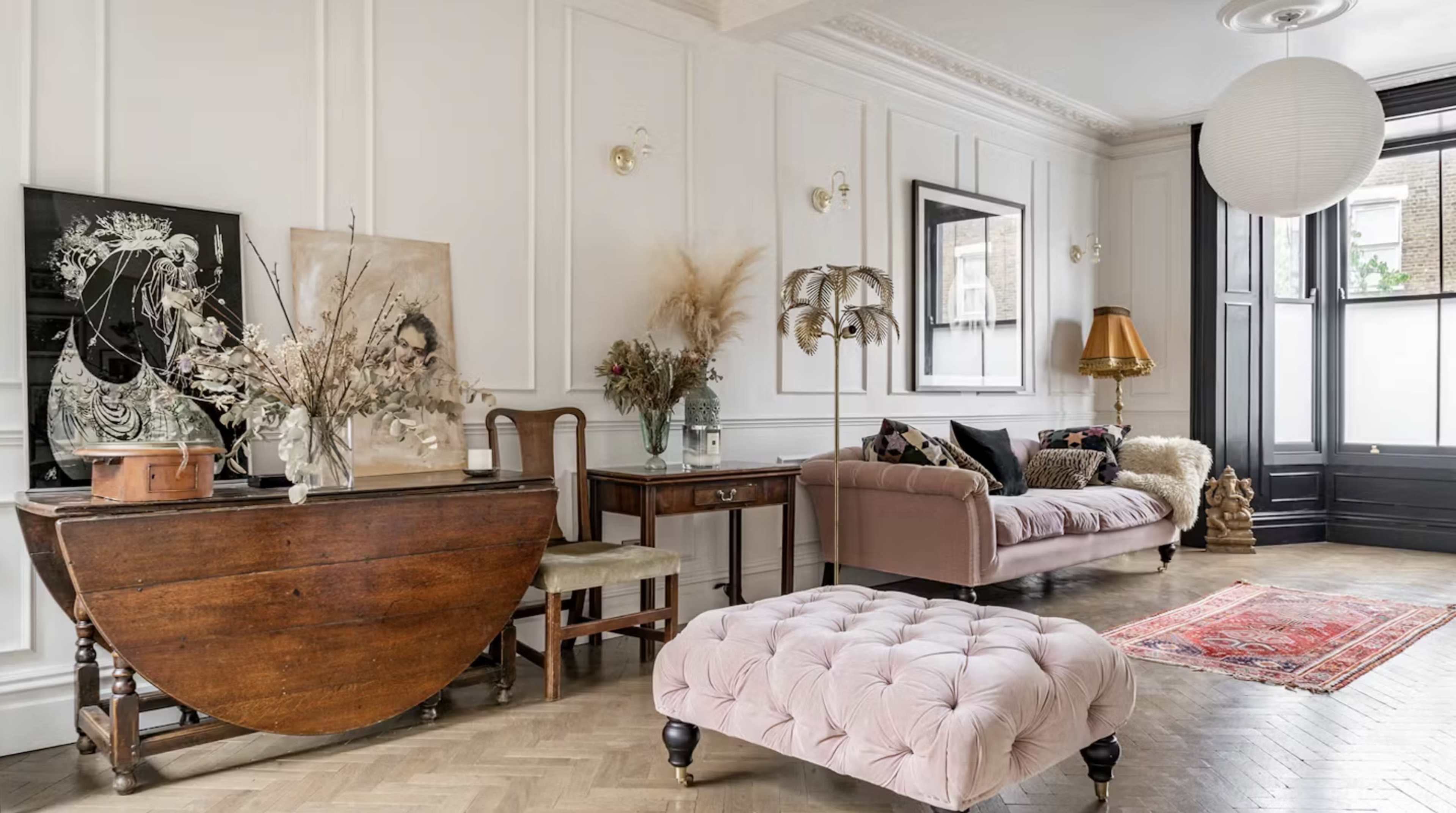 The room features a blend of vintage and modern furniture, including a pink tufted ottoman, a wooden table, and decorative plants against a backdrop of paneled white walls.