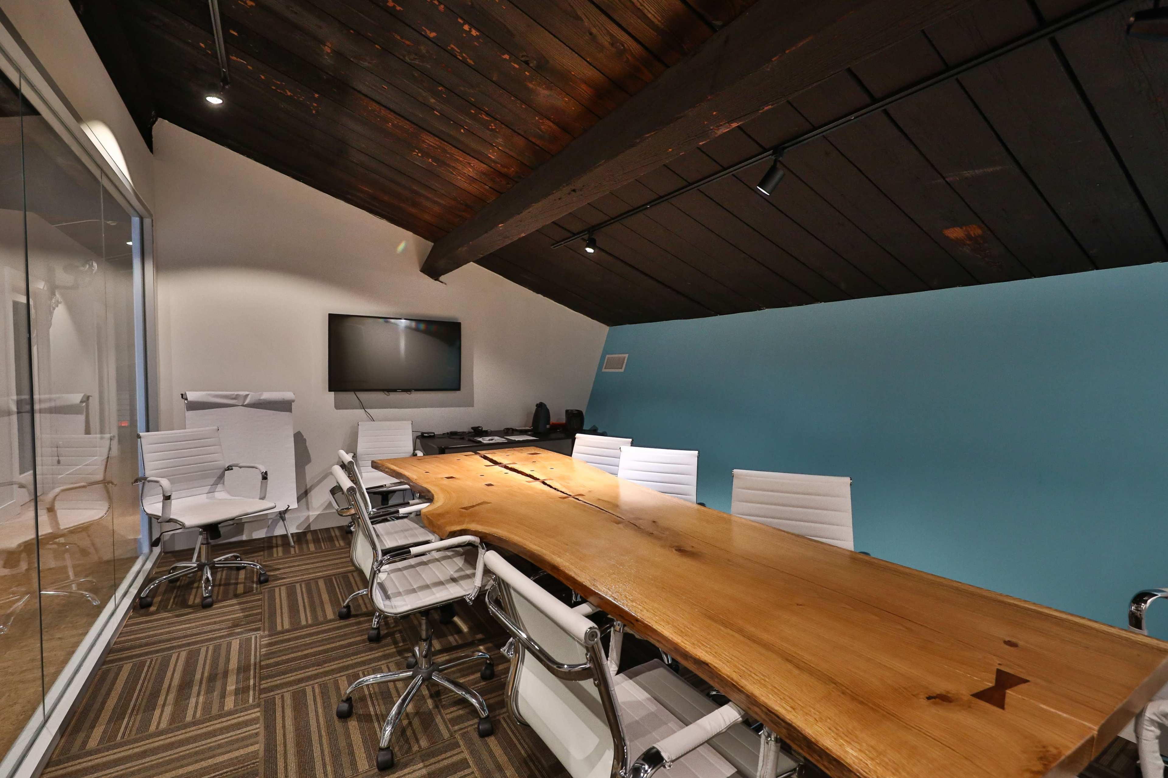 A modern conference room features a large wooden table surrounded by white chairs, with a blue accent wall and a television mounted on the wall.