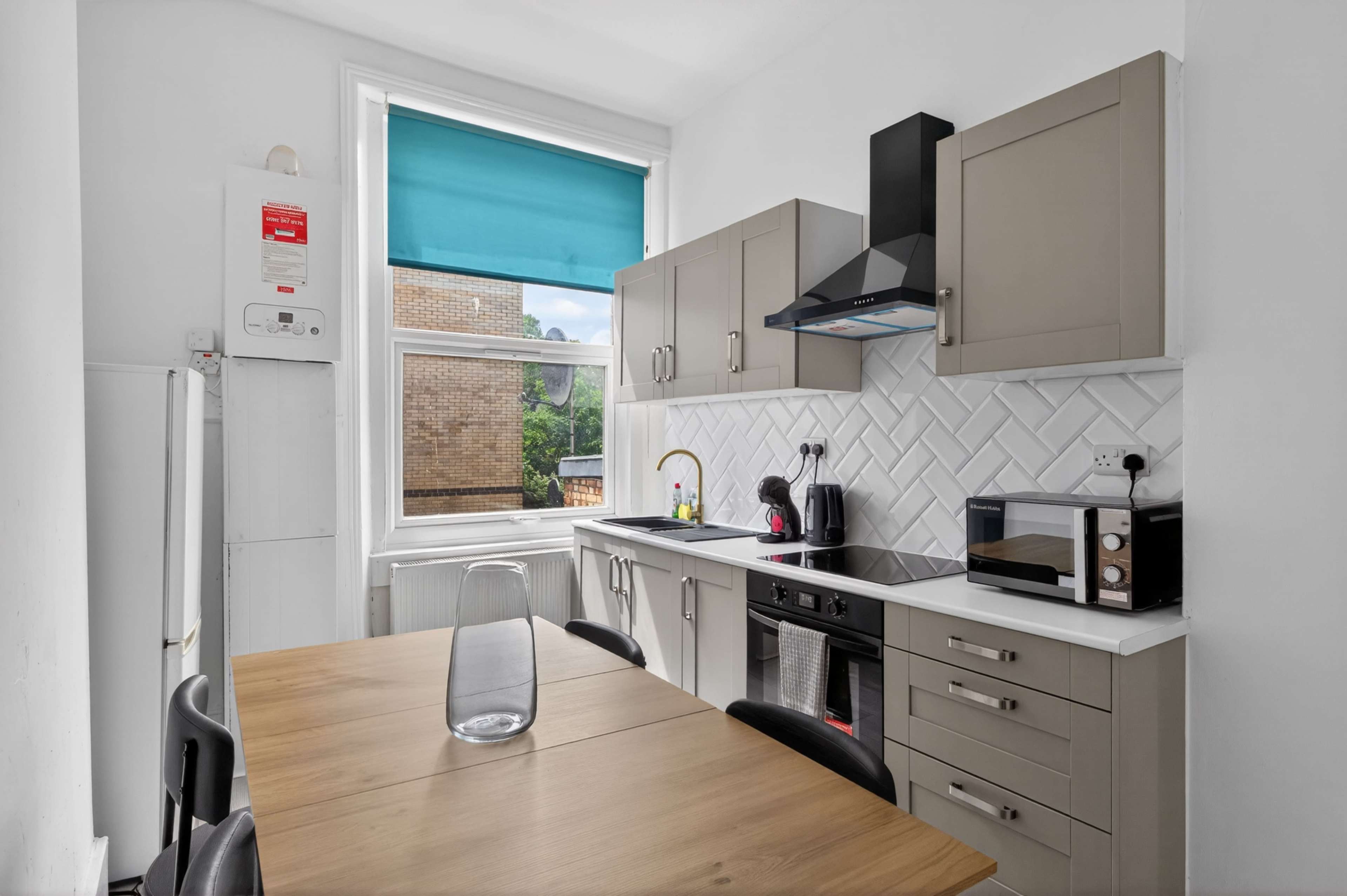 A modern kitchen features a wooden dining table, dark chairs, and contemporary appliances, with a large window covered by a turquoise roller blind.