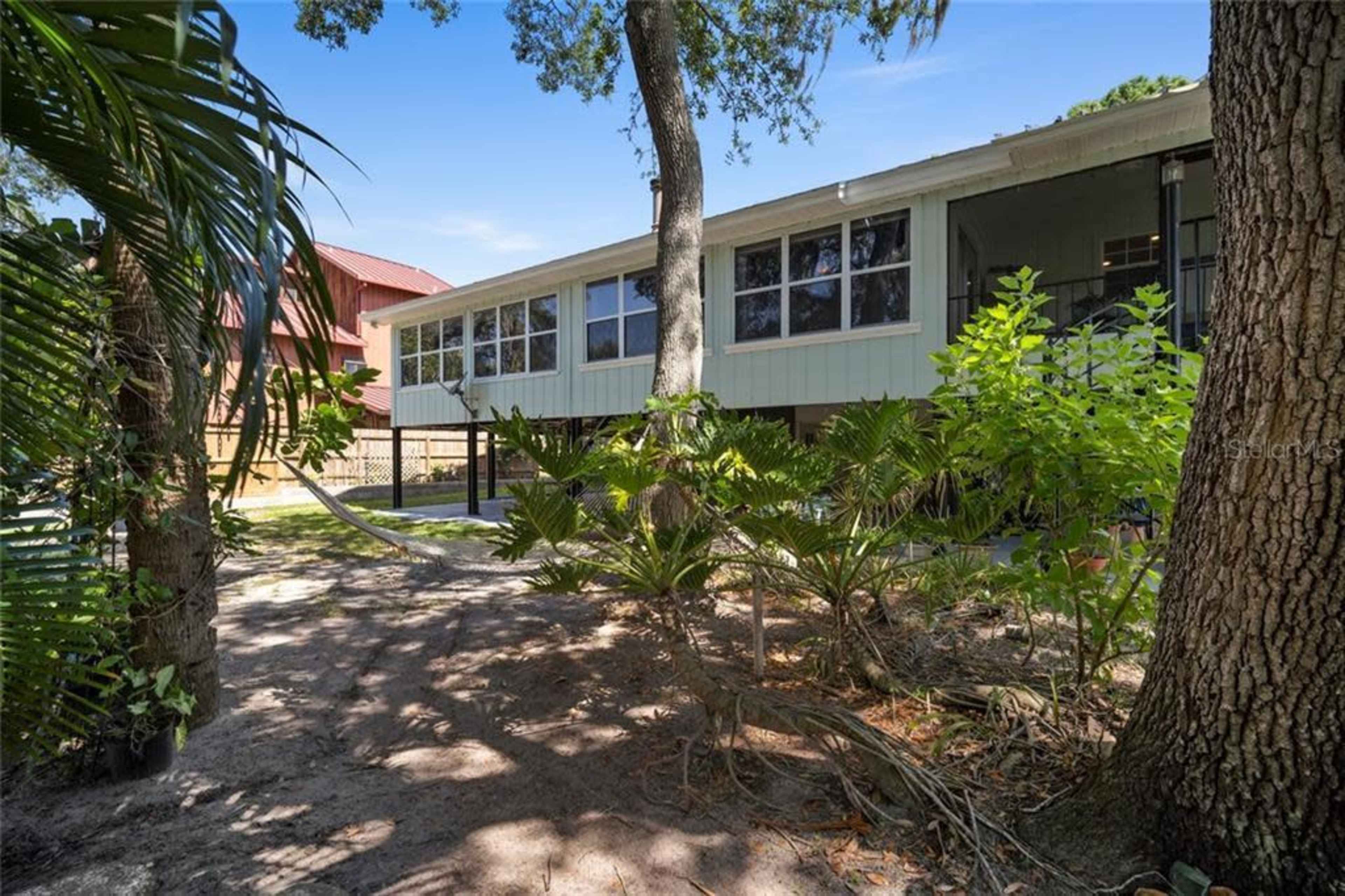 A house with a raised structure is surrounded by trees and tropical plants, with a sandy area beneath it.