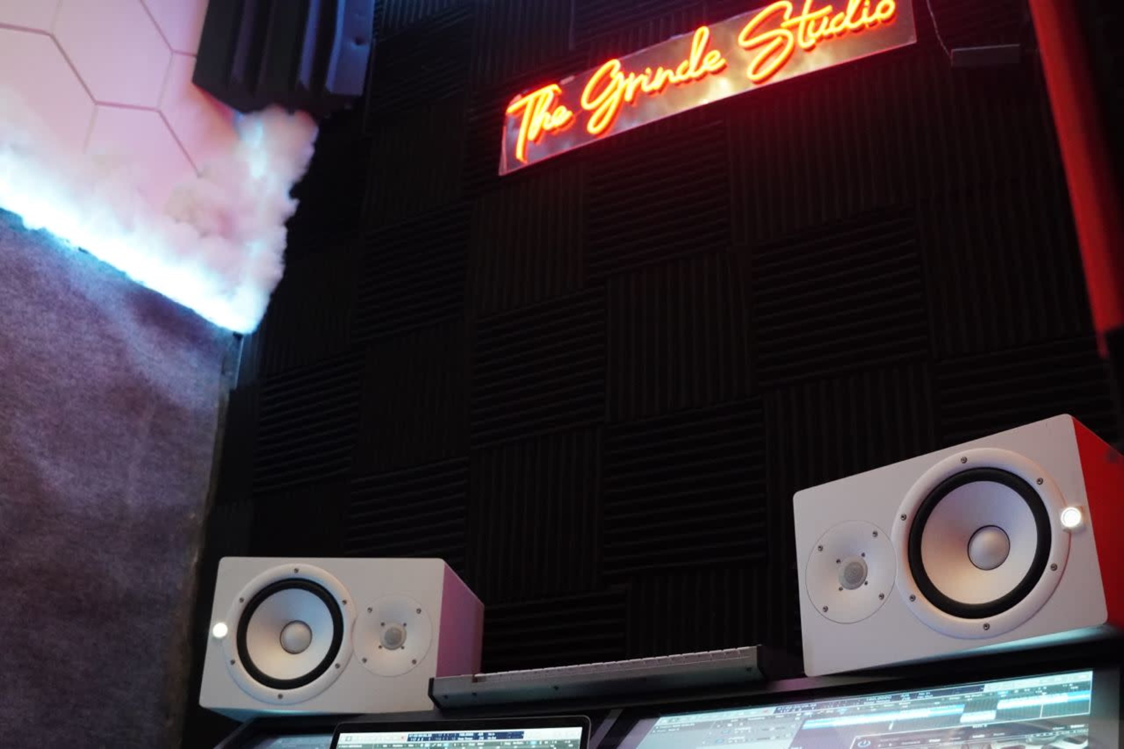 A sound studio with a neon sign reading "The Grind Studios" and two white studio monitors positioned on a desk.