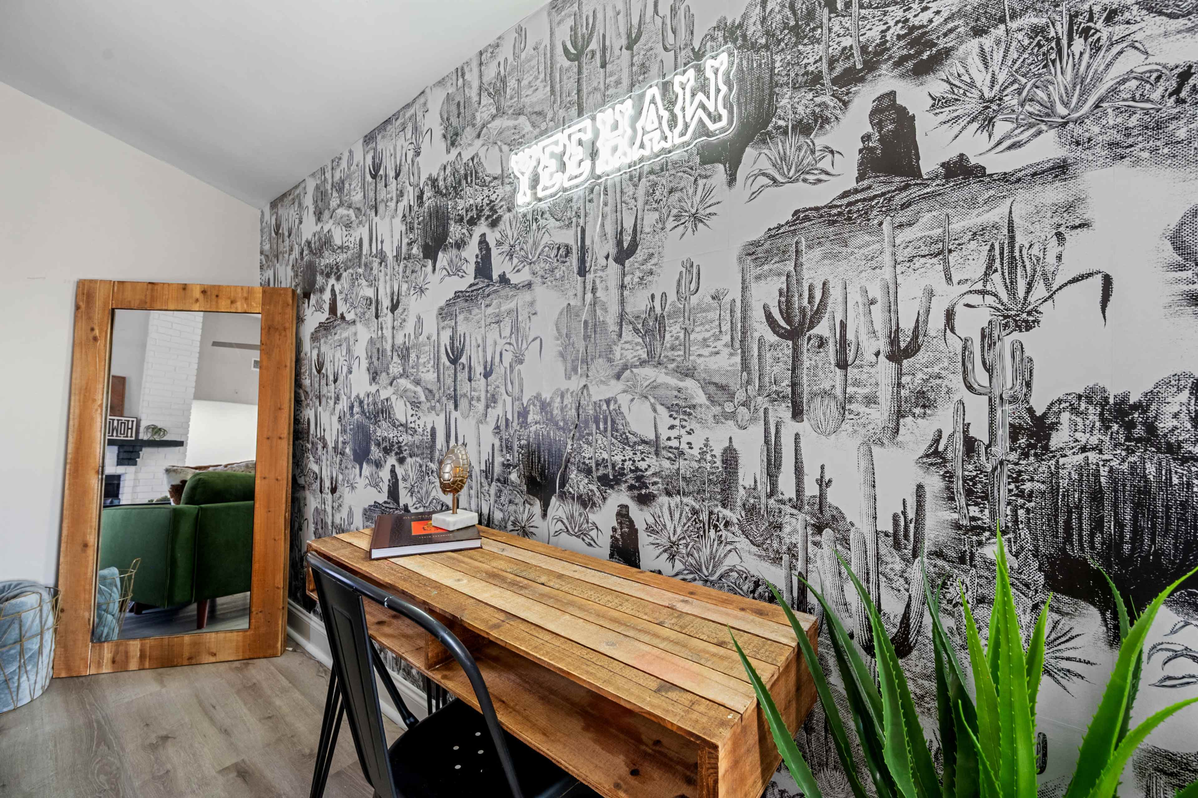 A wooden table sits in front of a cactus-themed wallpaper, with a mirror and a potted plant in the background.