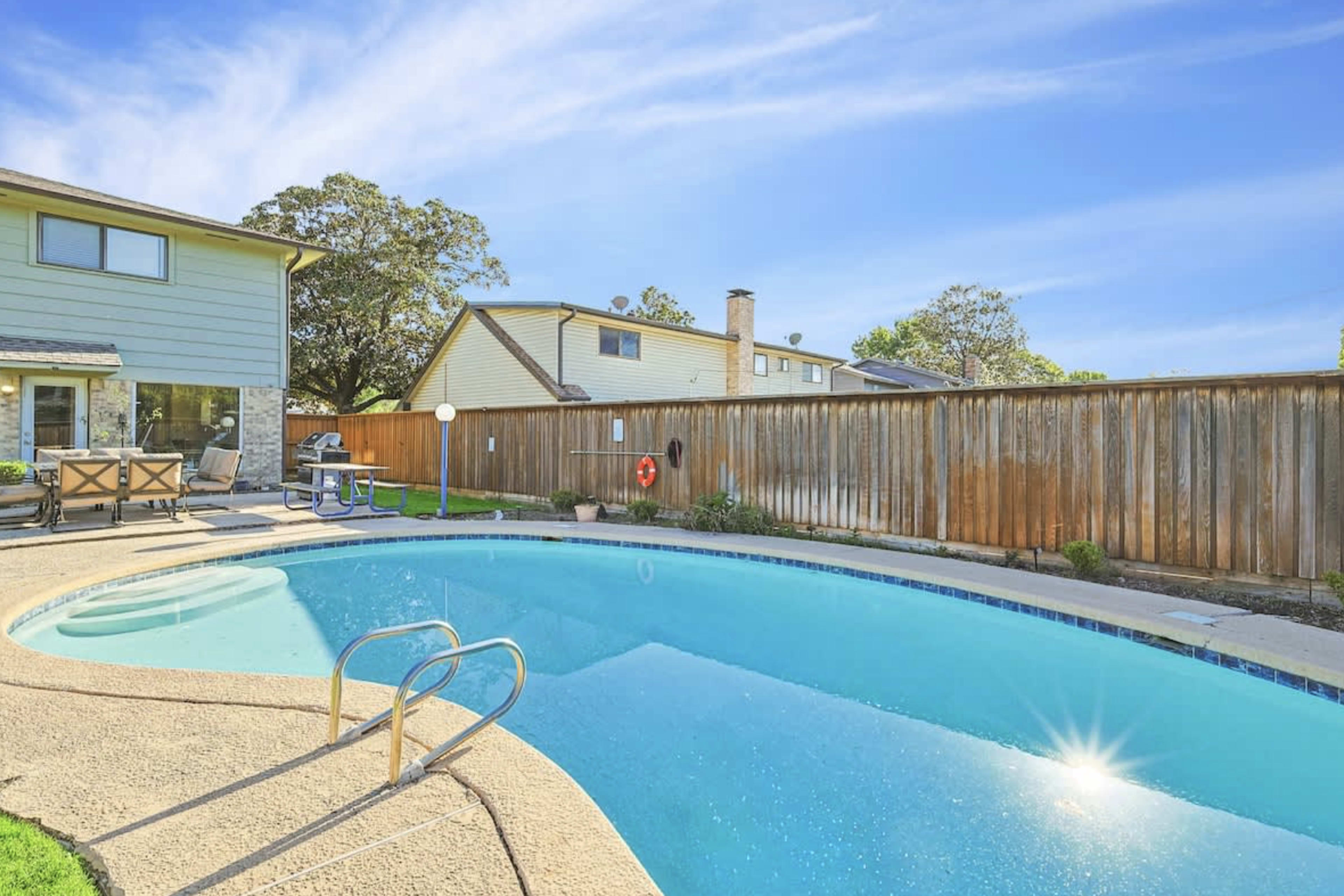 A clear swimming pool is surrounded by a concrete deck and a wooden fence, with a two-story house in the background.