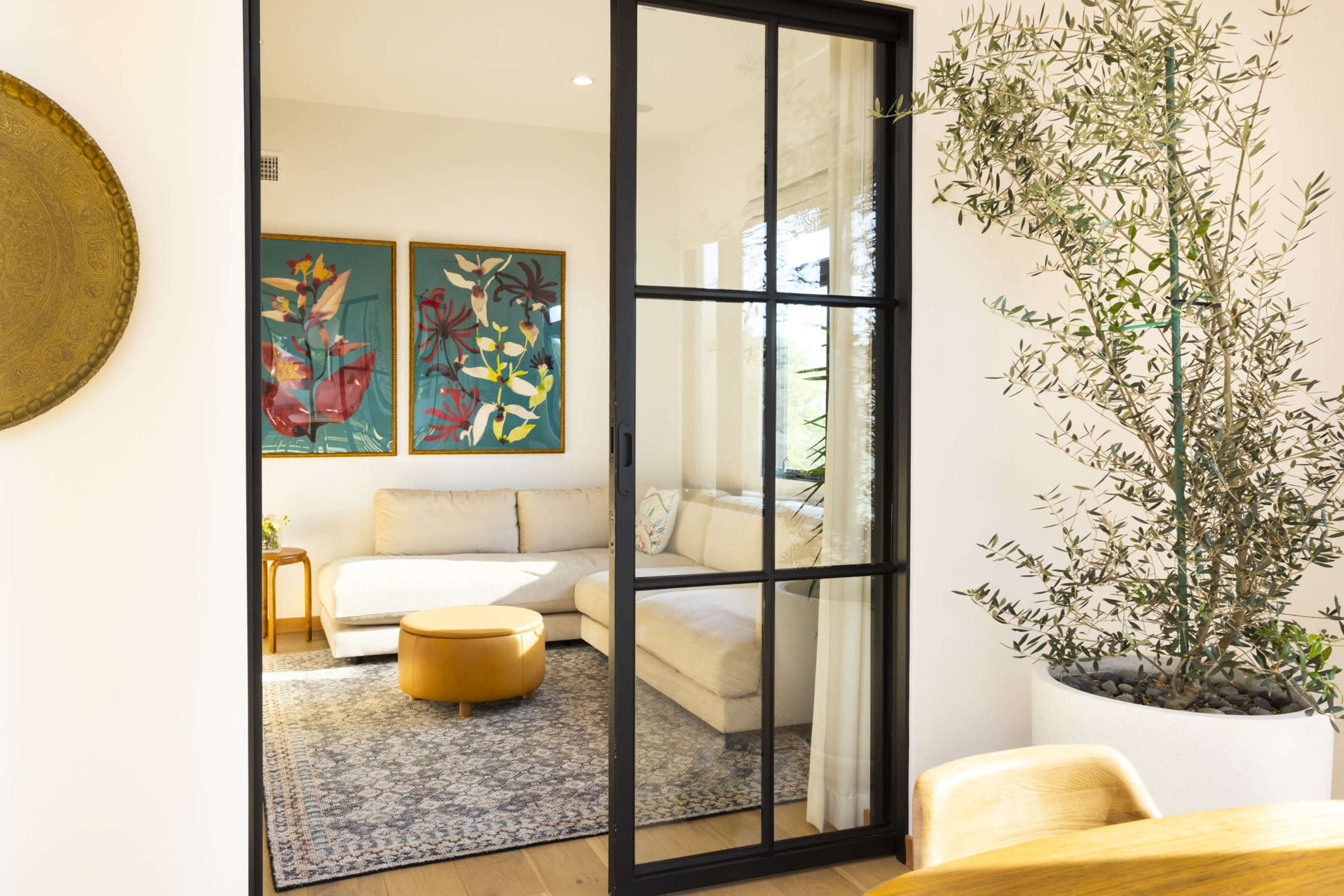 A modern living area features a light sofa, colorful artwork, and a decorative olive tree in a stylish pot.