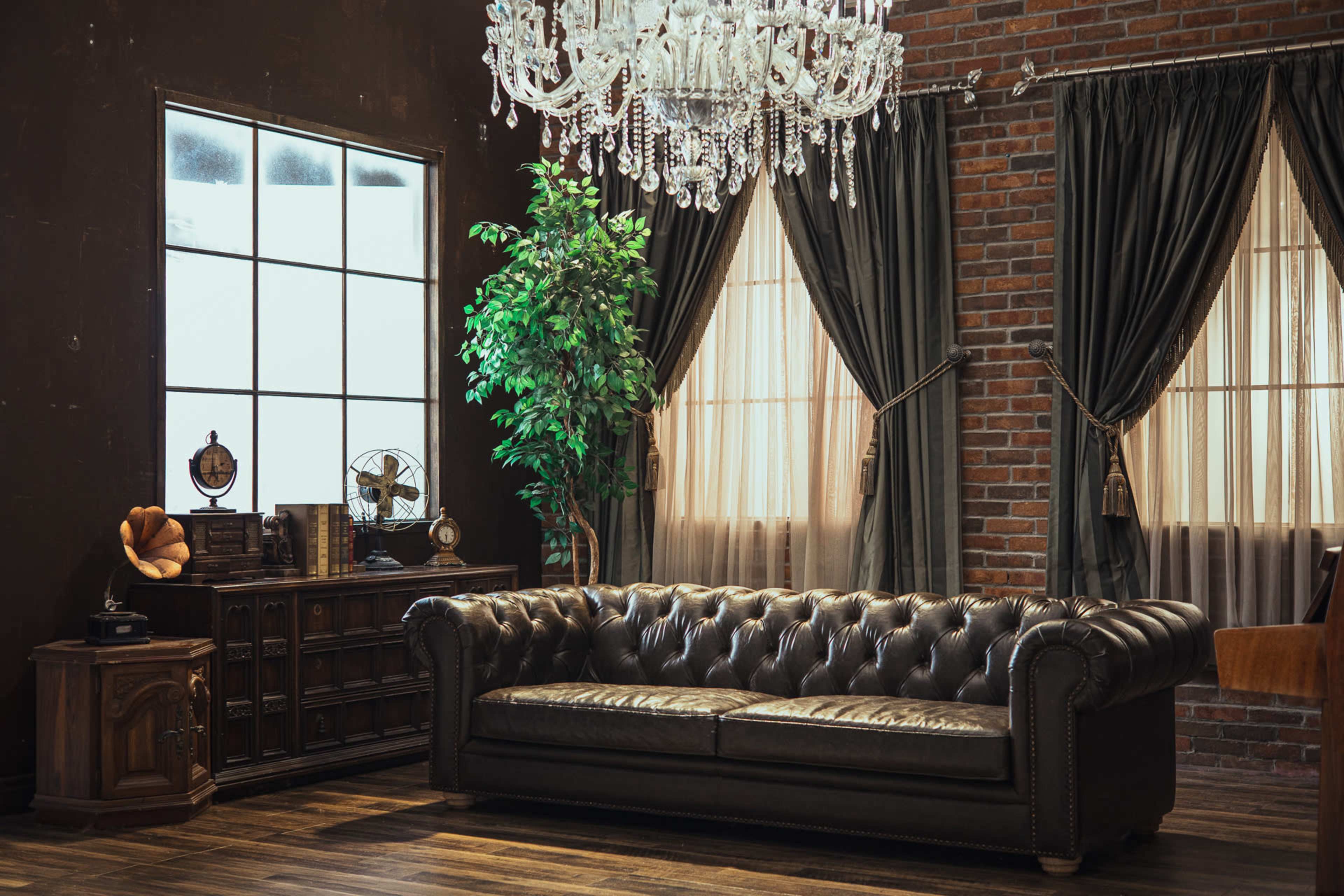The image features a dark, elegant room with a brown leather Chesterfield sofa, a large chandelier, brick walls, and large windows draped with heavy curtains.