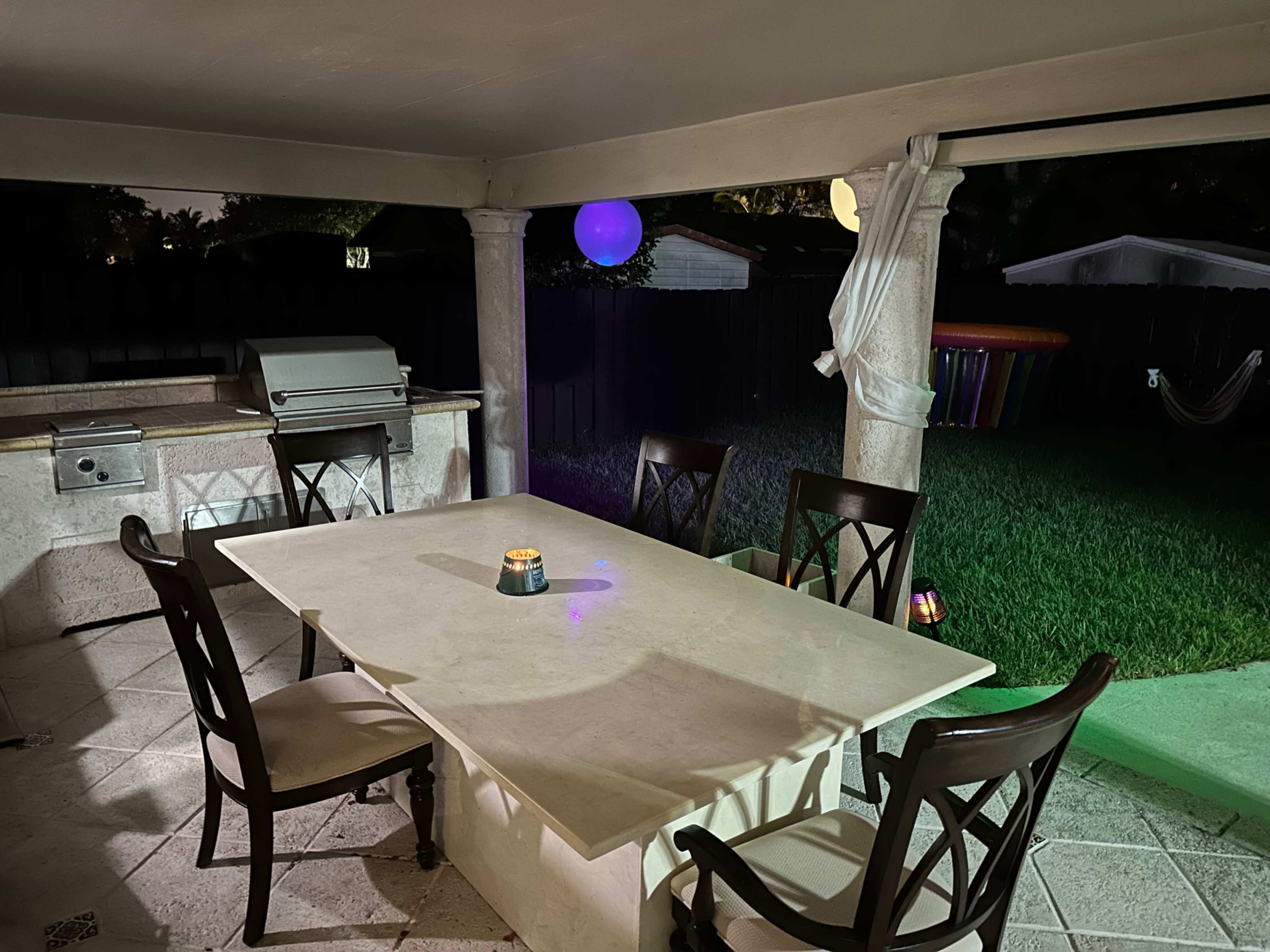 A covered outdoor dining area features a marble table surrounded by chairs, with a barbecue grill and ambient lighting in a grassy backyard.