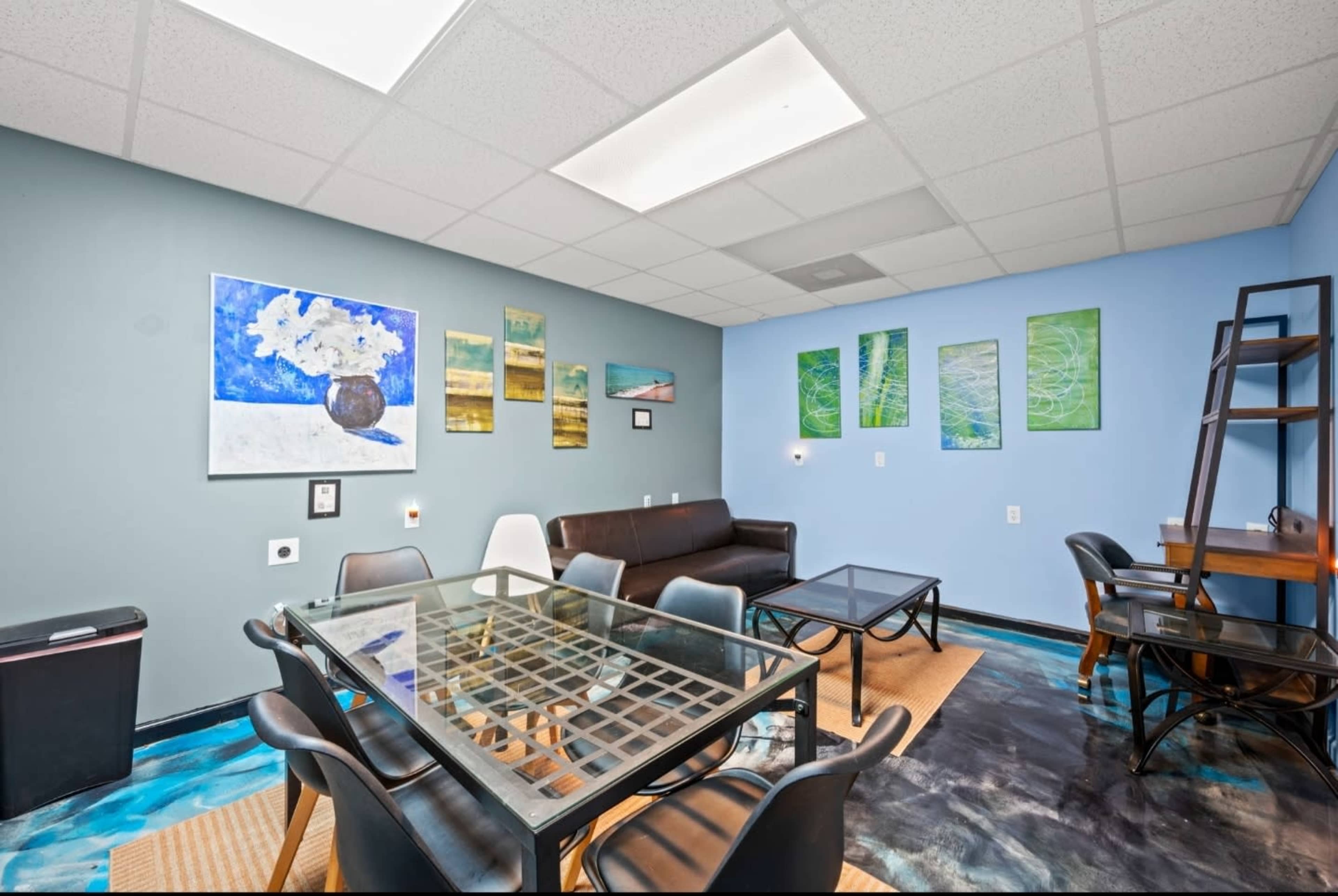 The image shows a modern meeting room featuring a glass table surrounded by black chairs, a couch, and colorful artwork on the walls.