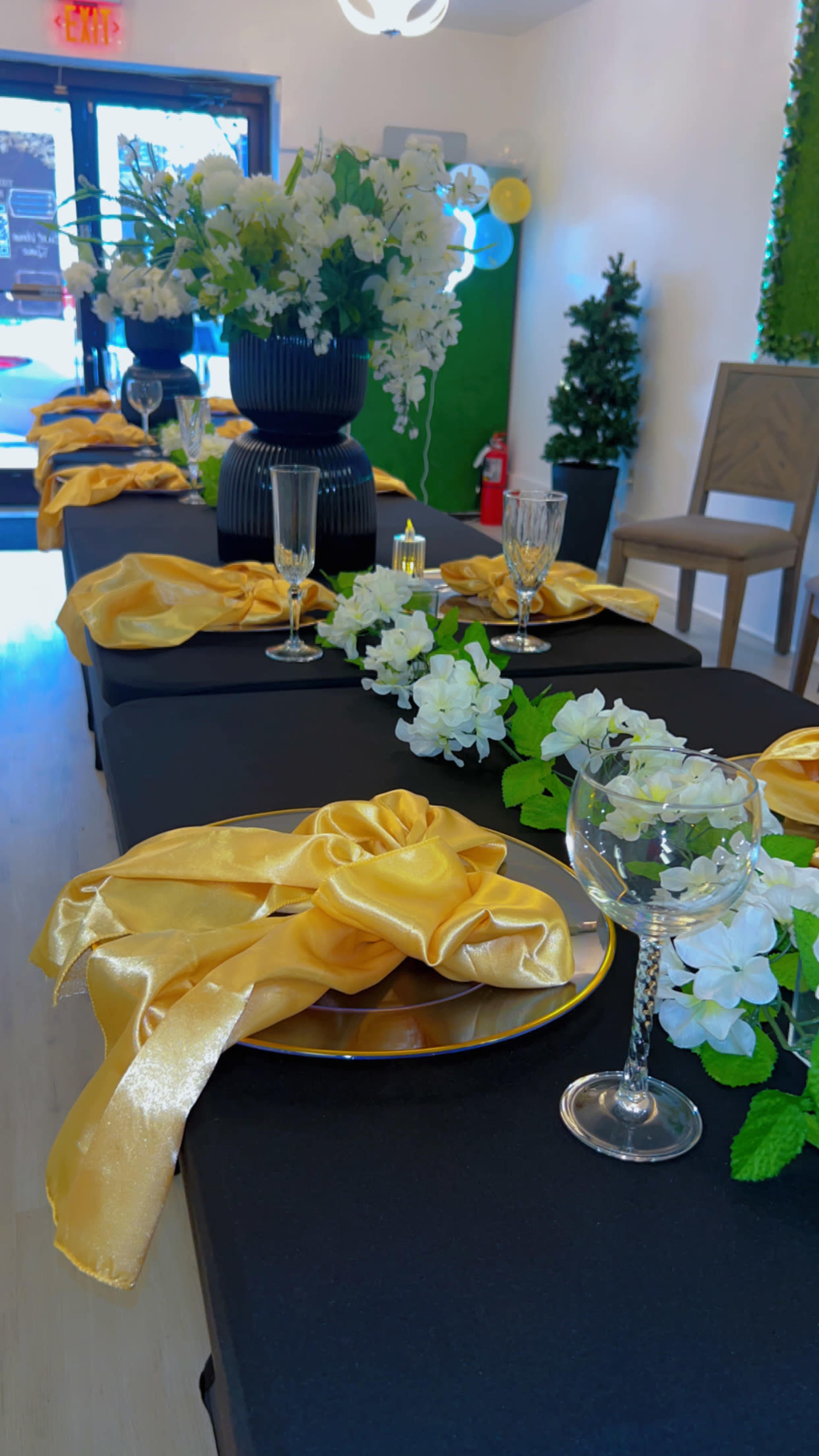 A neatly arranged table setting features black tablecloths, yellow satin sashes, glassware, and floral decorations.
