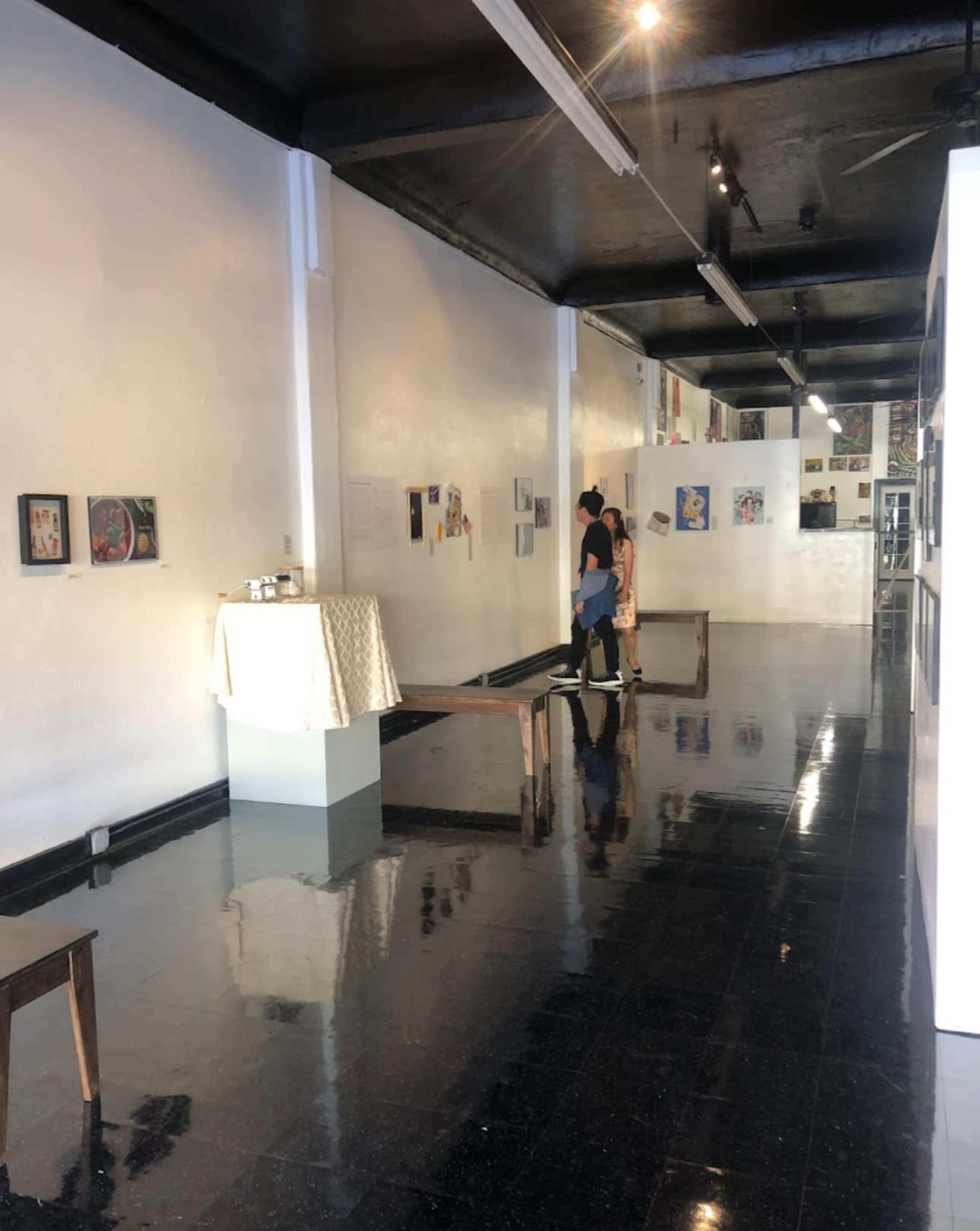 Two people are exploring an art gallery with white walls, black floors, and various artworks displayed.