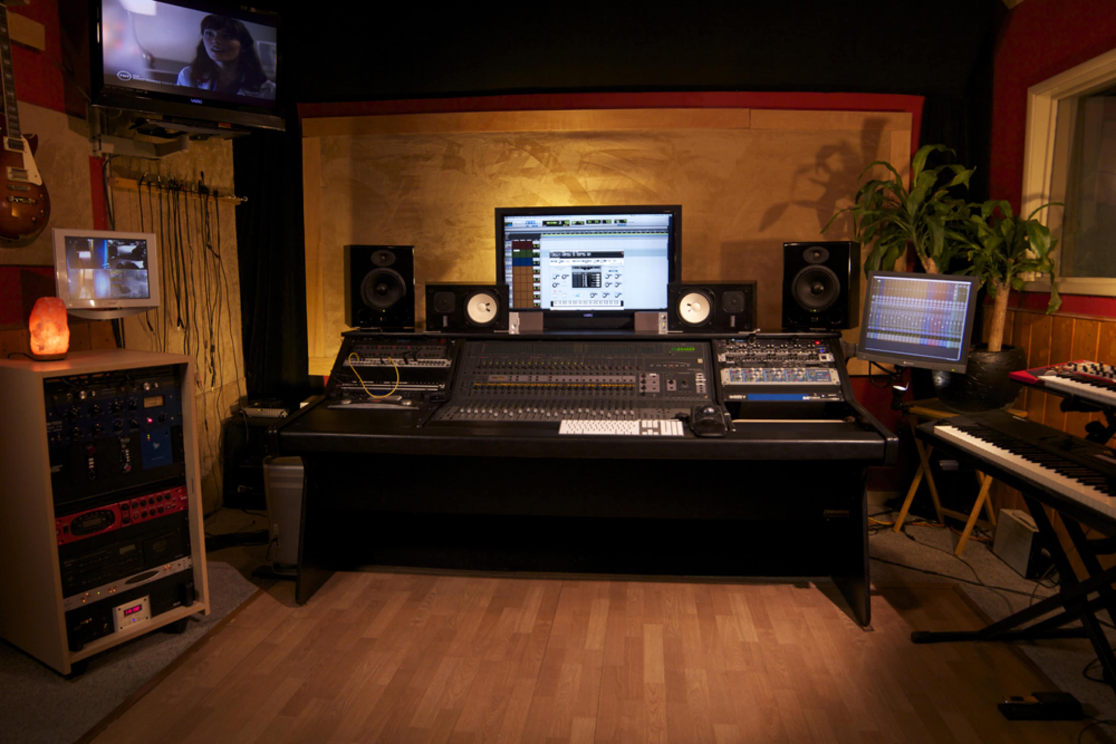 The image shows a music recording studio featuring a large mixing console, multiple monitors, audio equipment, and musical instruments.