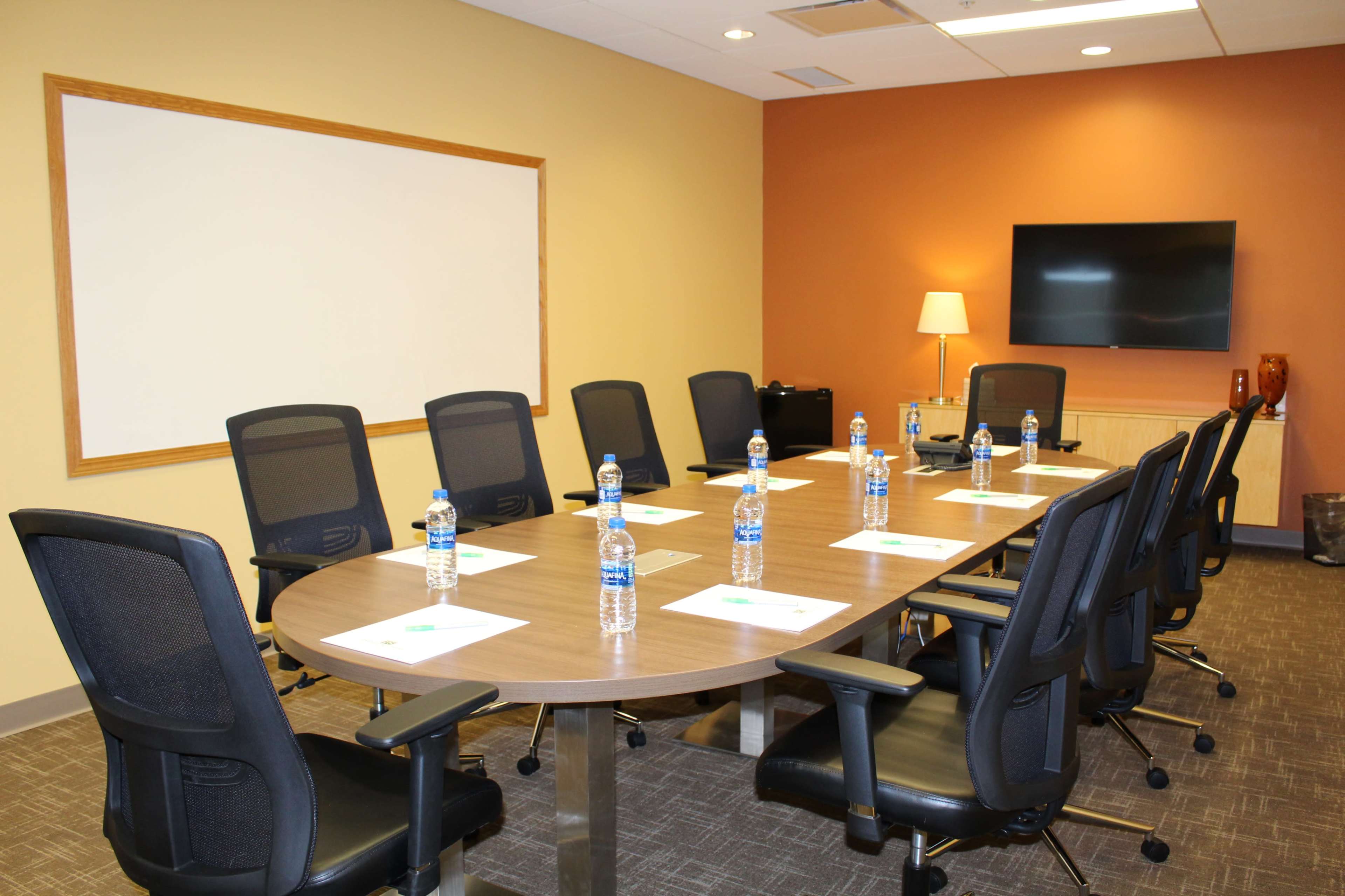 A conference room is set up with a large oval table surrounded by black chairs, each equipped with a water bottle and a notepad, along with a television and a whiteboard on the walls.