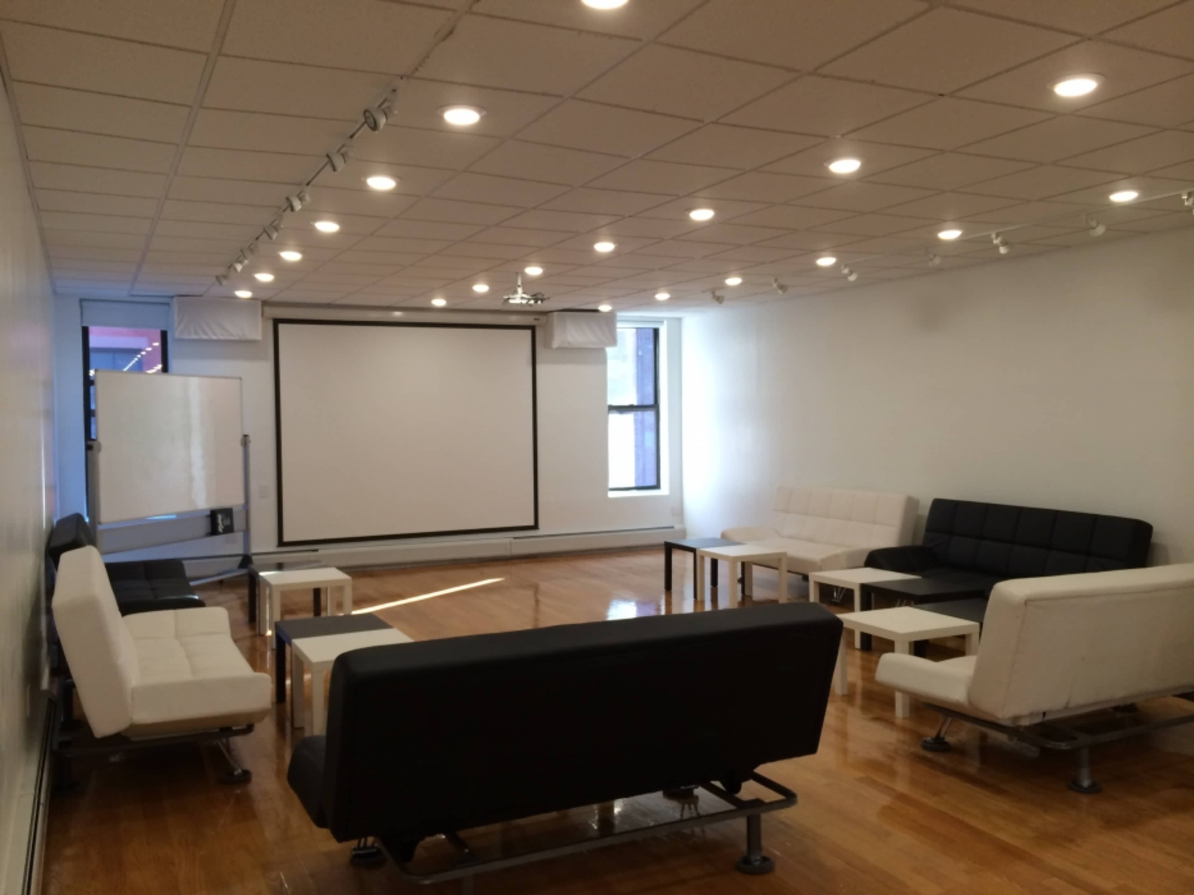 The room features a large screen at the front with several white and black sofas arranged around tables on a polished wooden floor.