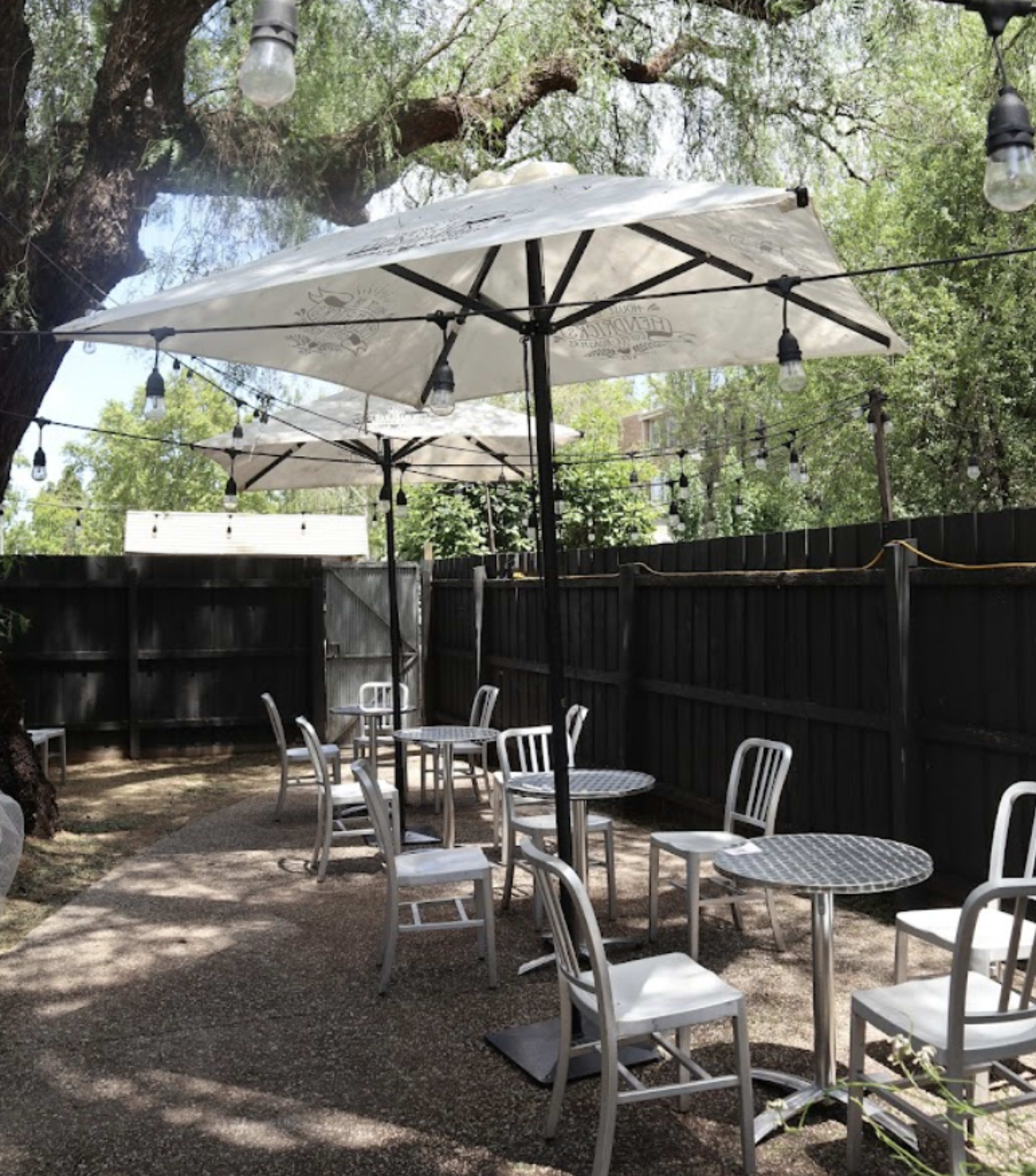 A patio area features several tables with white umbrellas surrounded by a wooden fence and string lights.