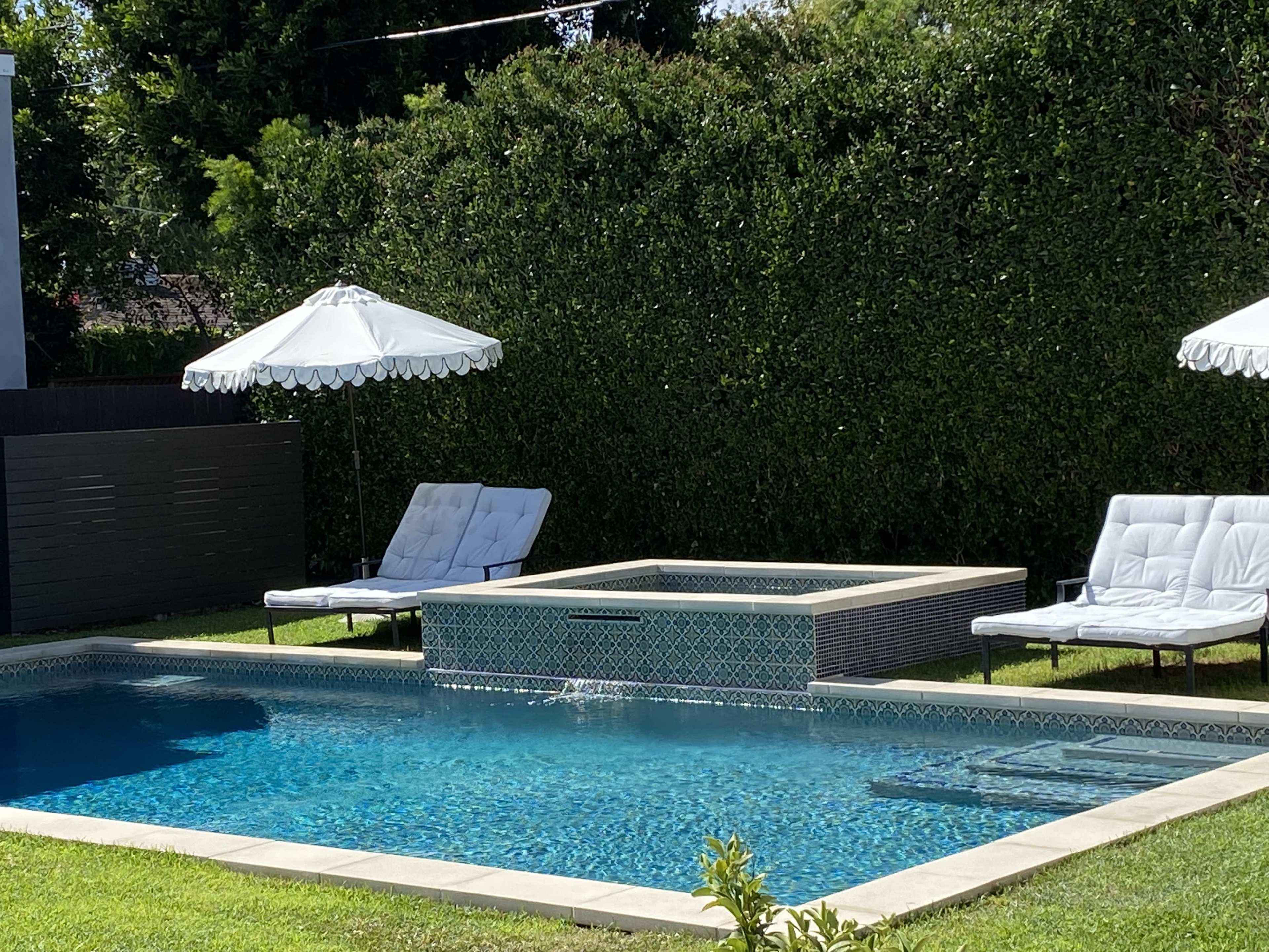 A rectangular swimming pool with a spa is surrounded by two lounge chairs and white umbrellas, set in a green backyard.
