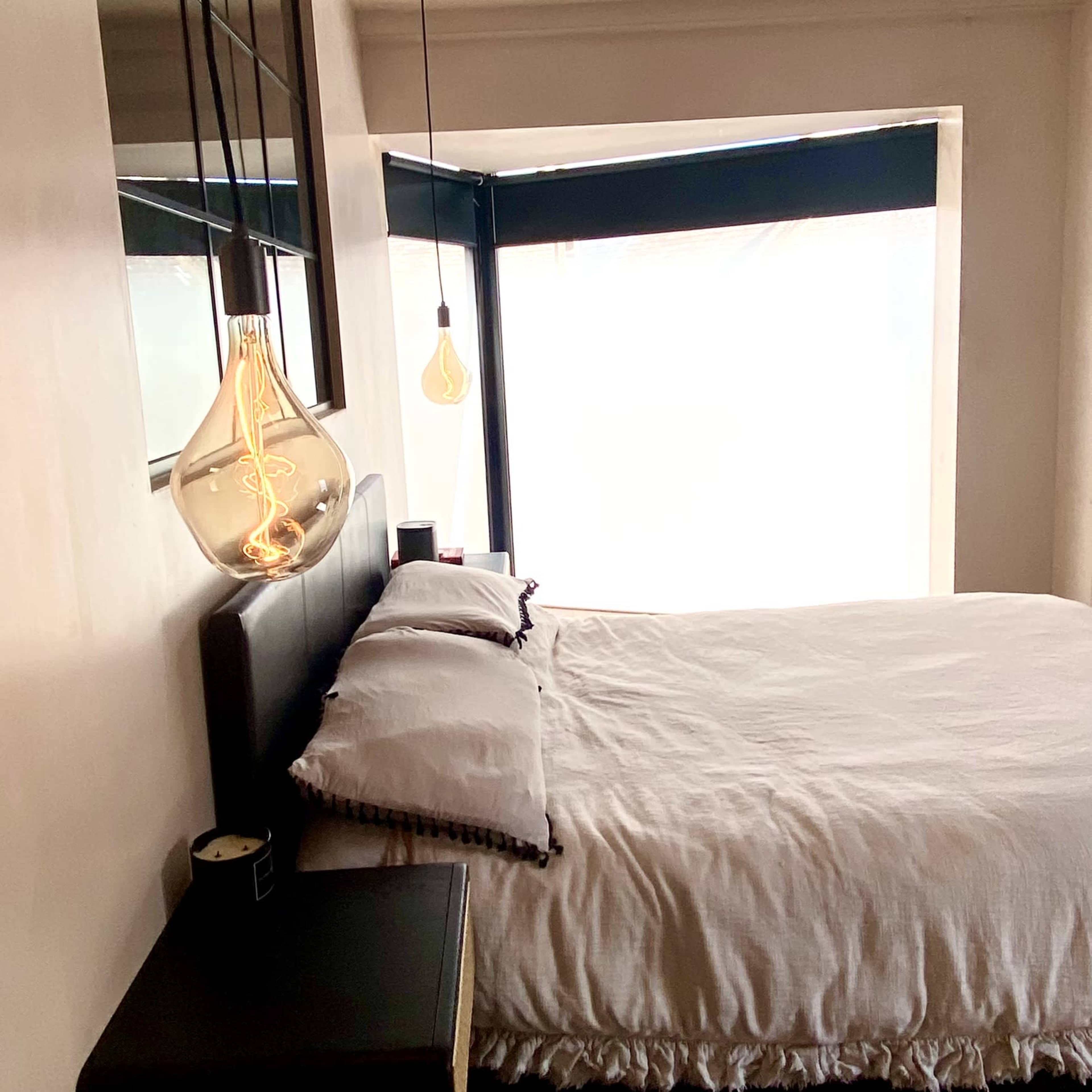 A well-lit bedroom features a bed with white linens, a small nightstand, and pendant lights hanging above.