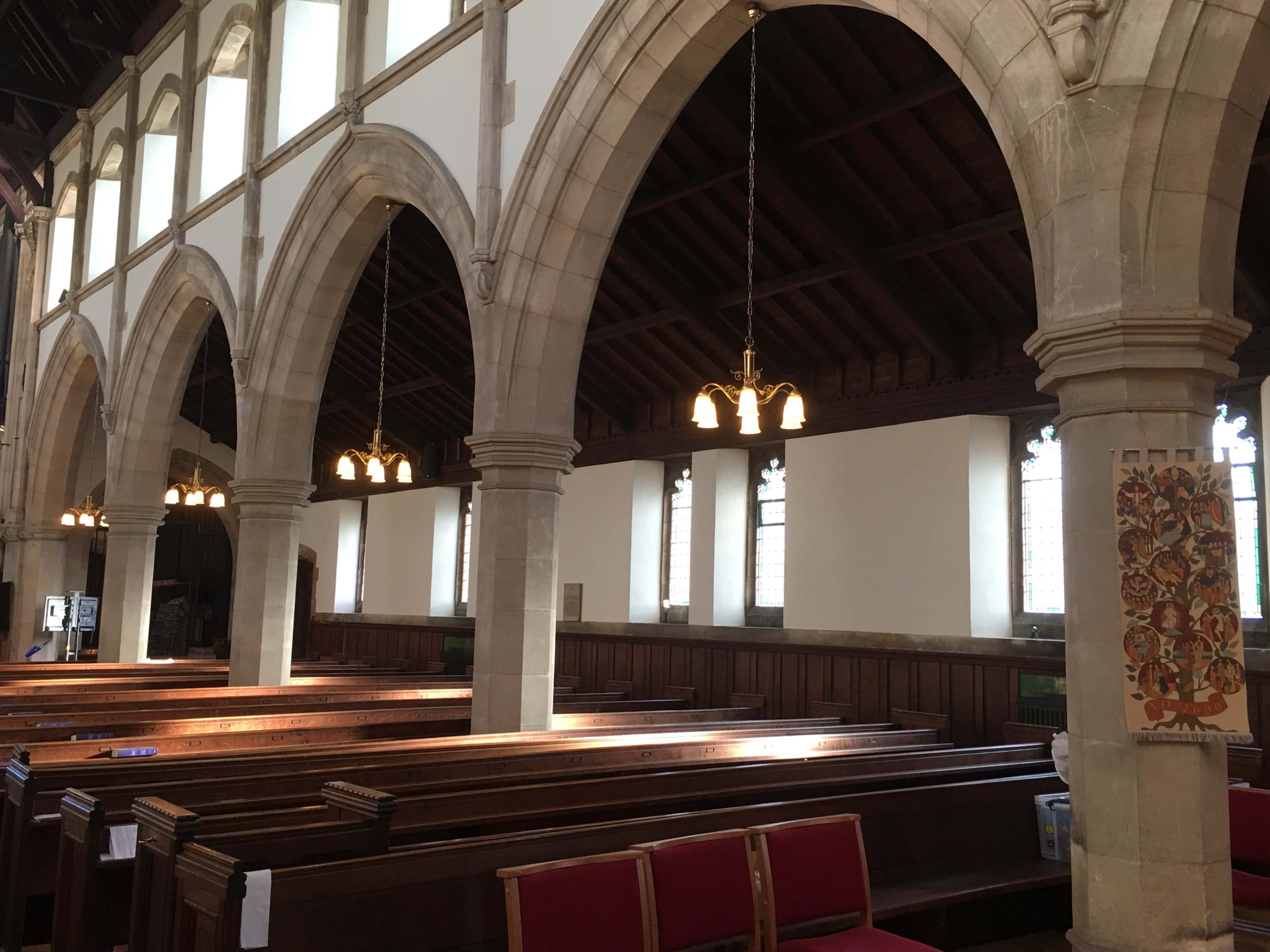 A church interior featuring wooden pews, arched stone columns, and large windows with a wooden ceiling above.