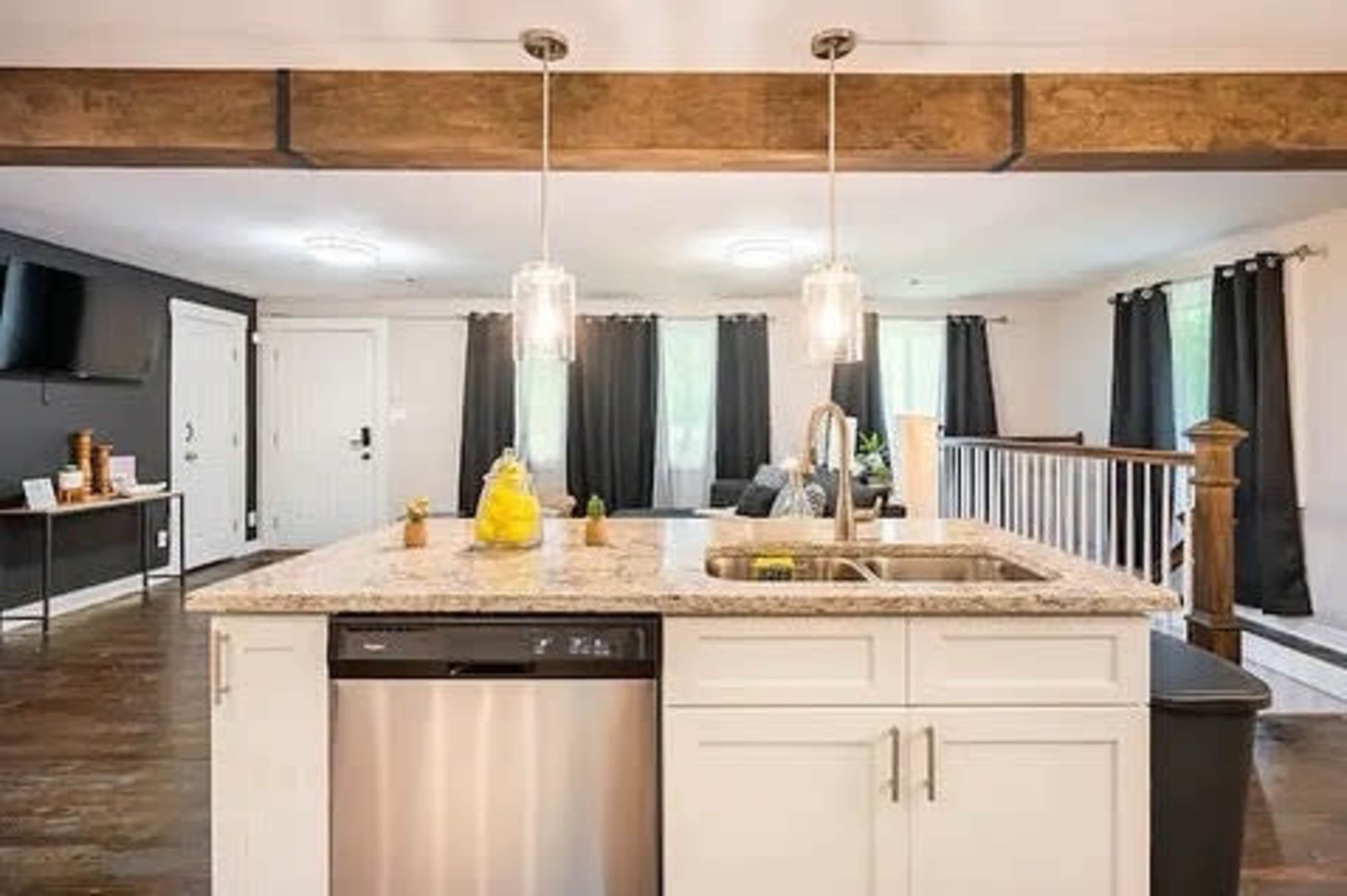 A modern kitchen island with a sink, dishwasher, and pendant lights, overlooking a living area with a sofa and large windows.