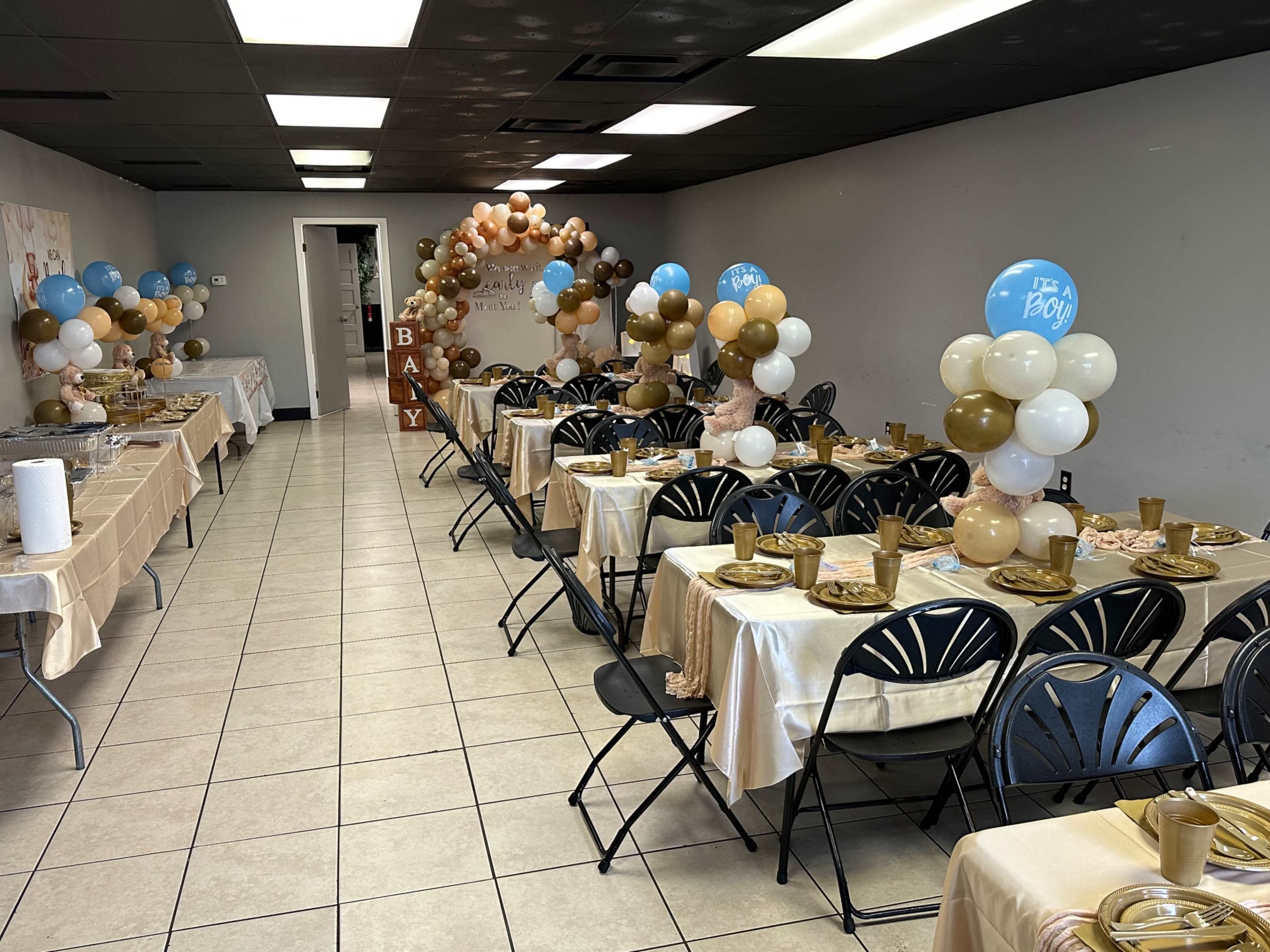 The image shows a decorated event space with tables set for a celebration, featuring gold and blue balloons, tableware, and themed decorations.