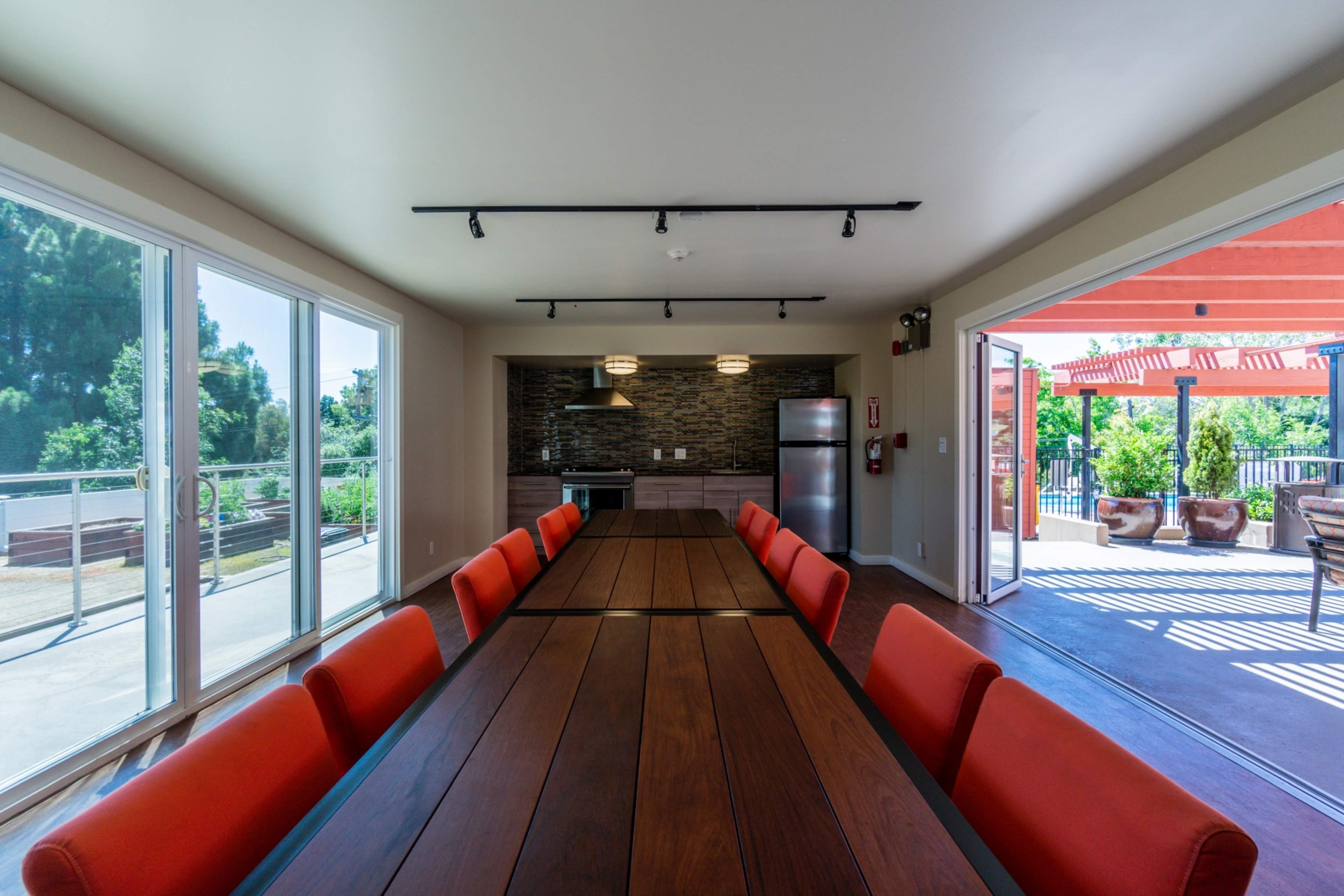 A long wooden table with orange chairs is positioned in a brightly lit room featuring large windows and a kitchenette area.