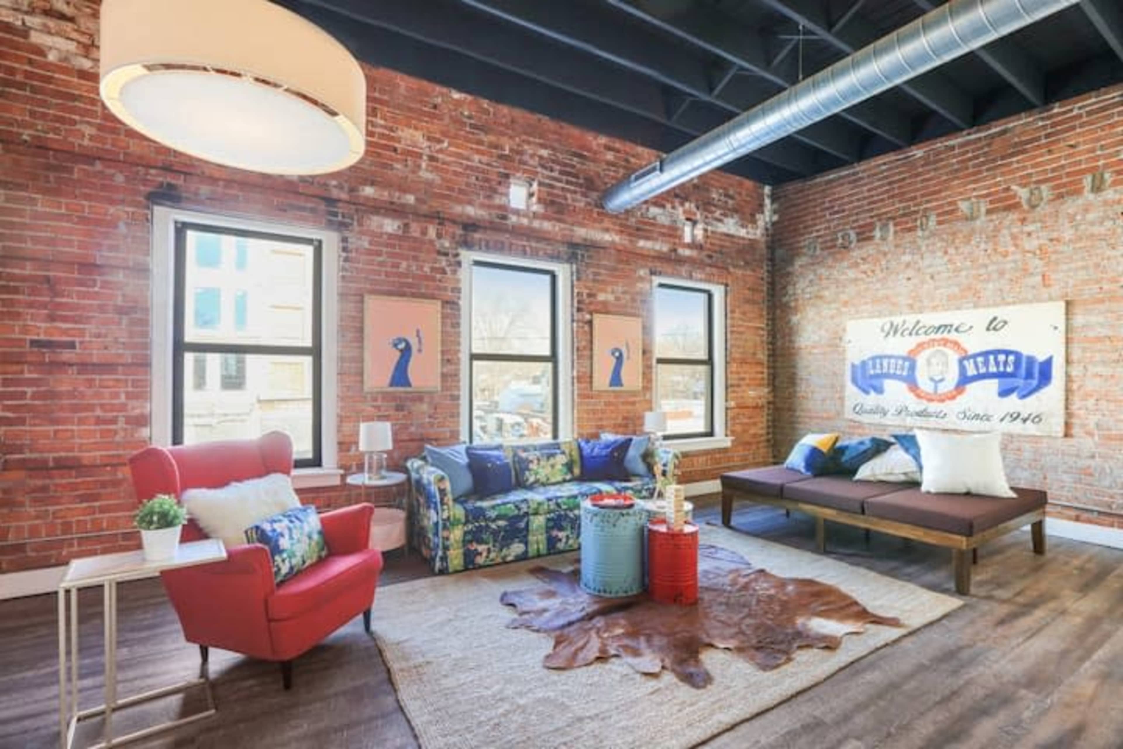 The living room features a mix of red and blue furniture, a large area rug, exposed brick walls, and large windows letting in natural light.