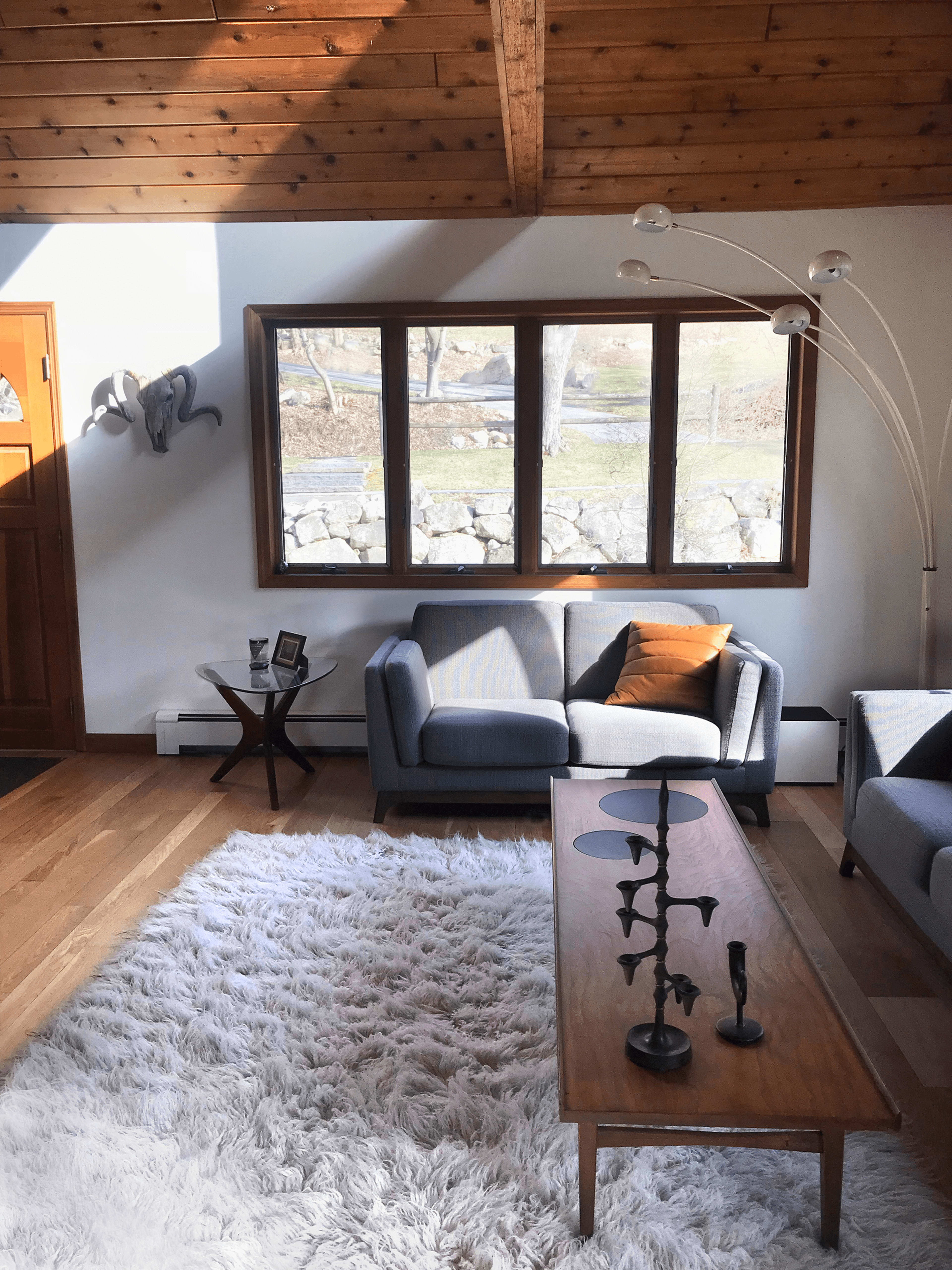 The image depicts a bright living room featuring two gray sofas, a wooden coffee table, a white rug, and large windows that overlook a stone wall and outdoor space.