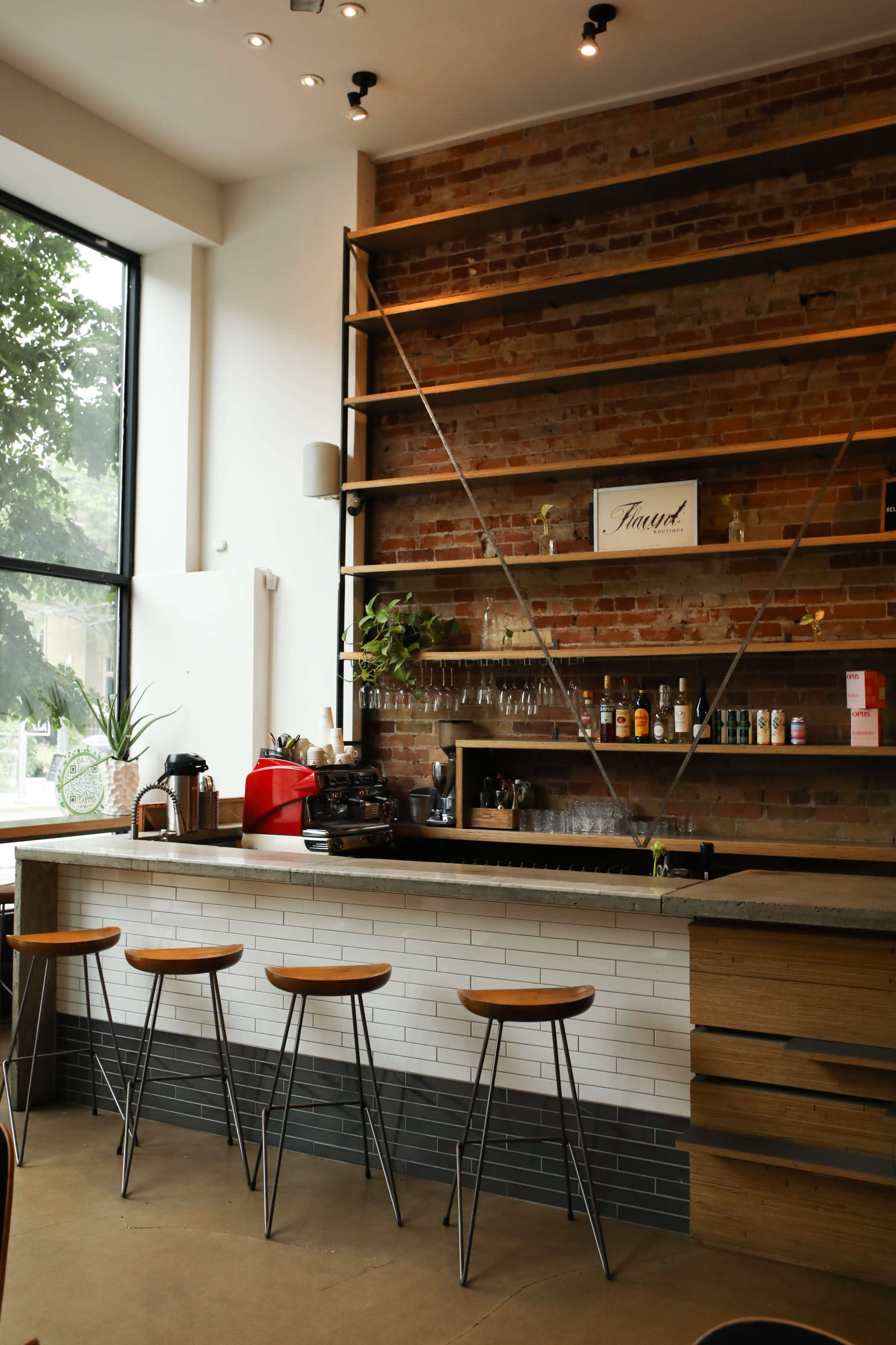 A modern coffee bar with a wooden counter, a brick wall, and four stool chairs in front of it.