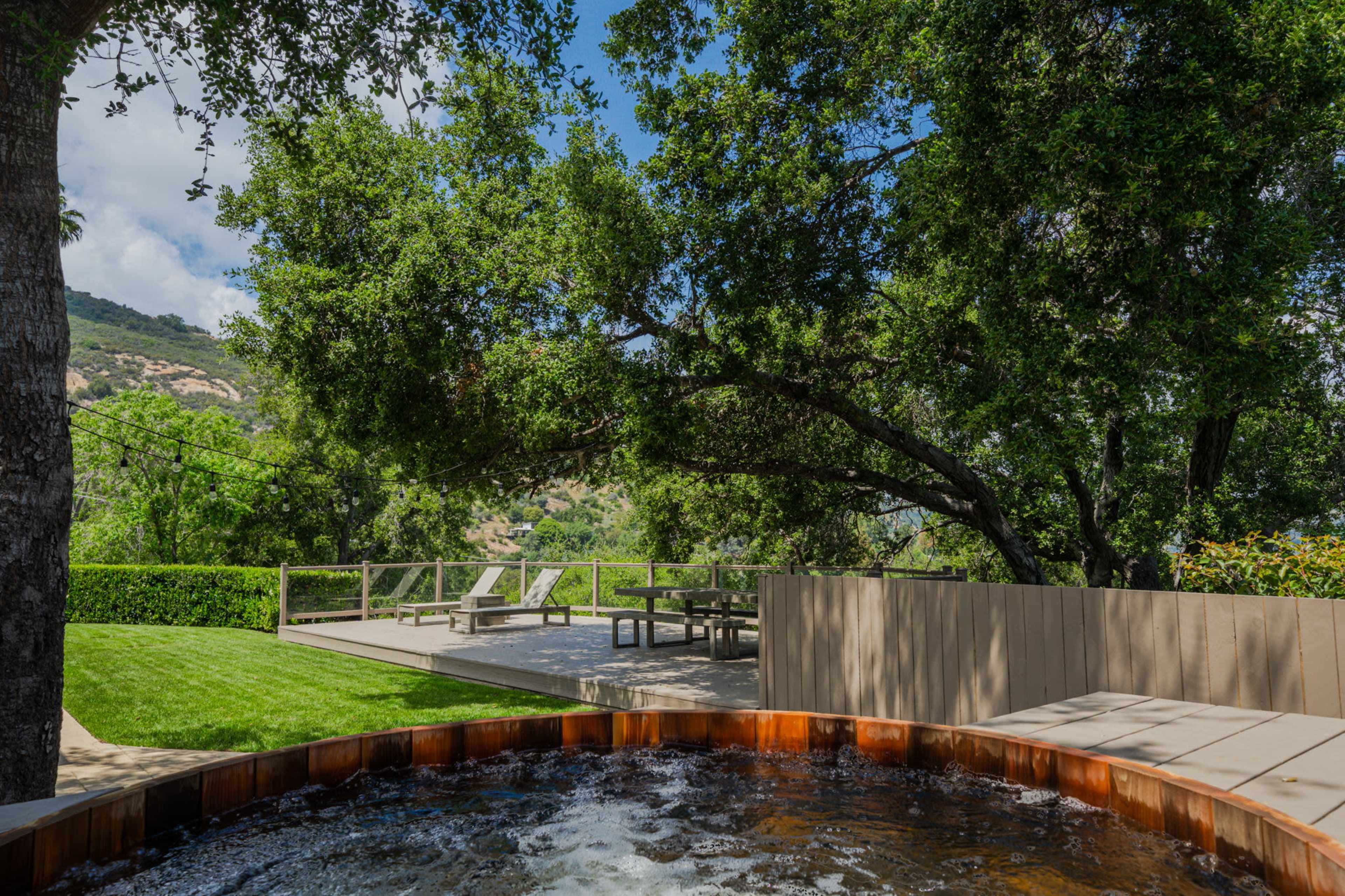 A hot tub sits in the foreground, surrounded by greenery, with a wooden deck and lounge chairs visible in the background under a sunny sky.