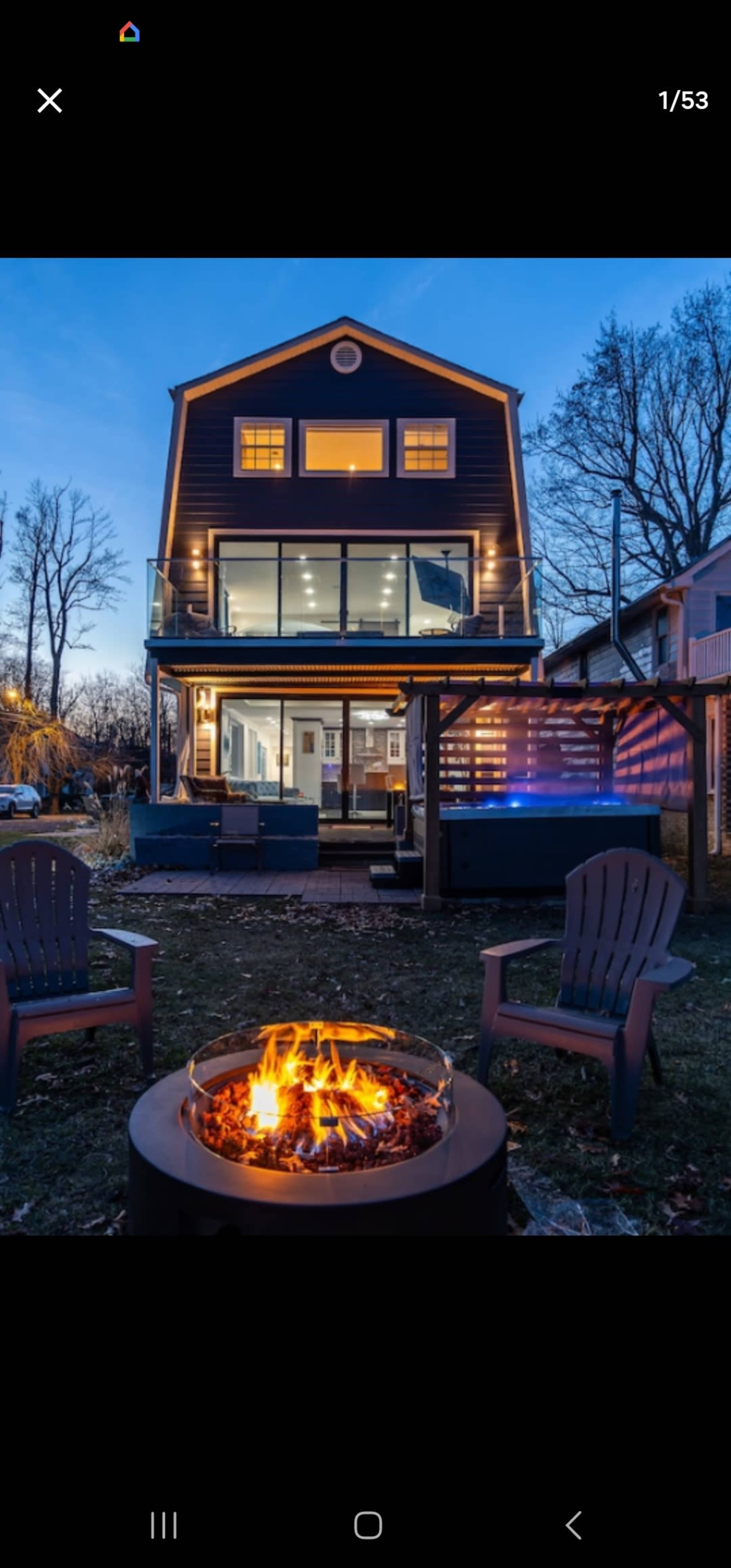 A modern two-story house with large windows and a deck is illuminated at dusk, with a fire pit and seating area in the foreground.