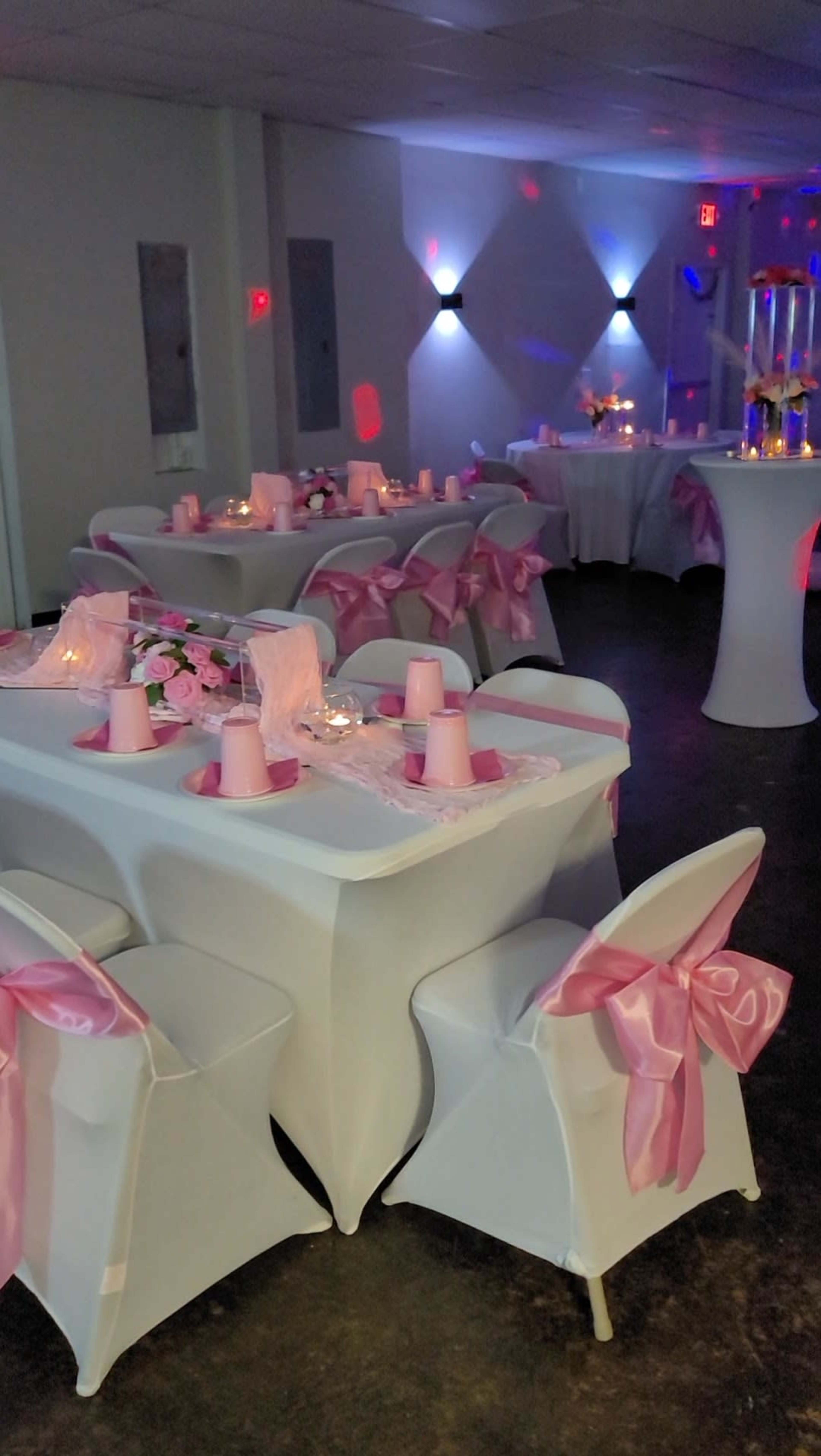 The scene shows a decorated event space with tables draped in white cloth and pink sashes, set with candles and floral arrangements.