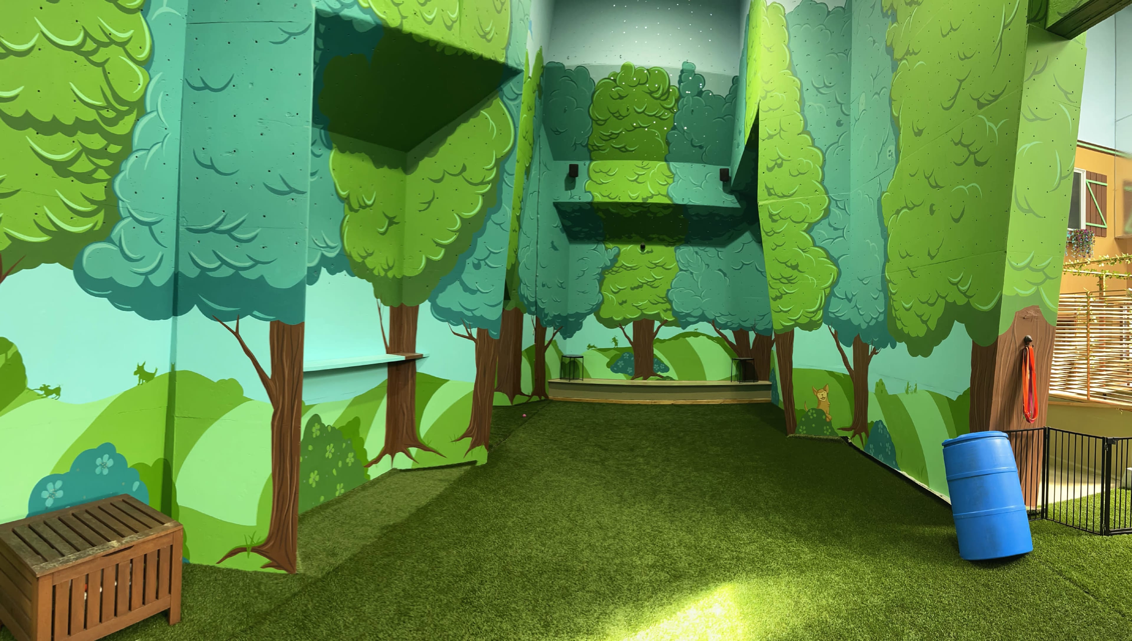 The image shows a brightly painted indoor space designed to resemble a forest, featuring large trees on the walls and artificial grass on the floor.