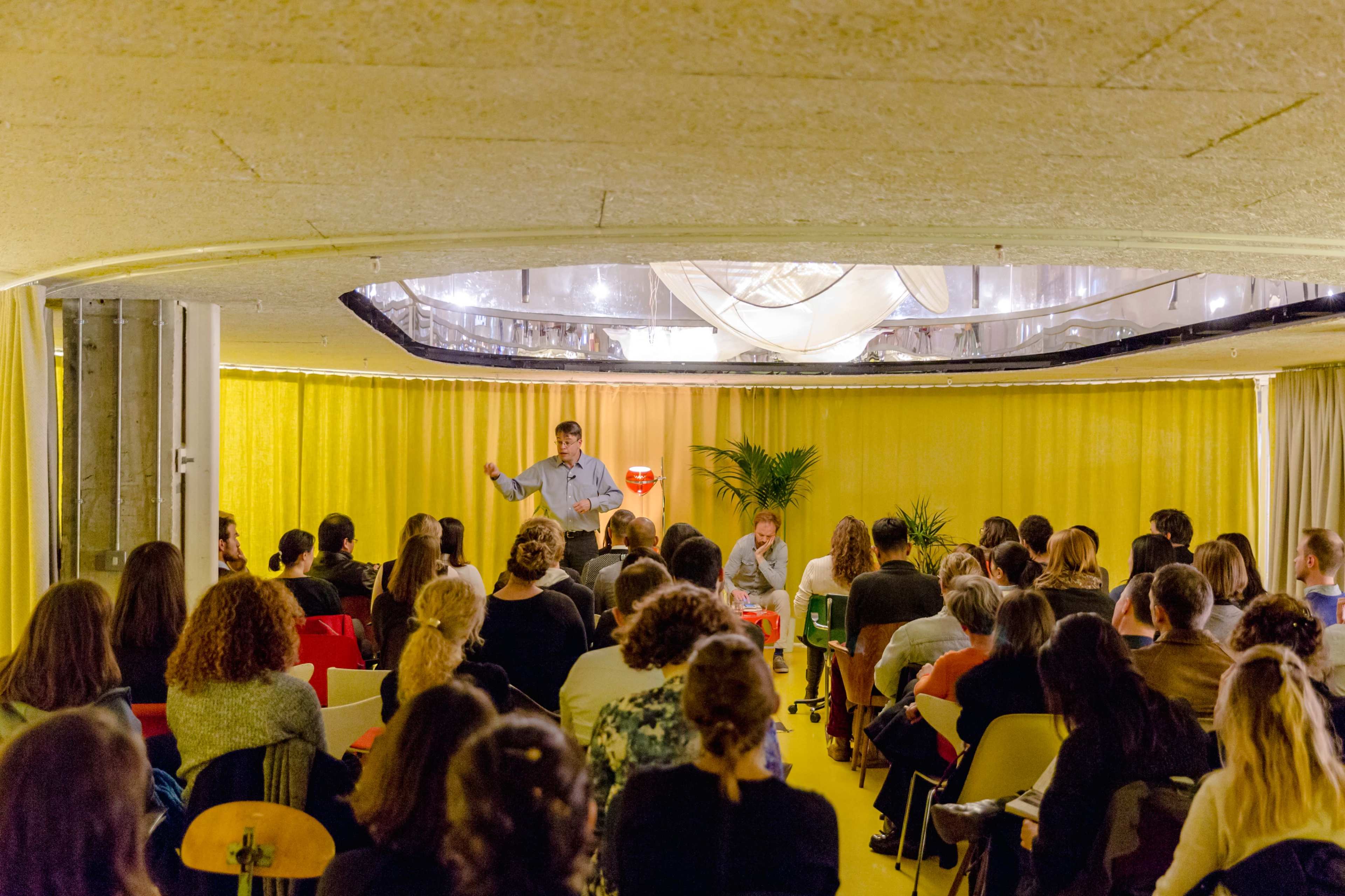 A speaker addresses a seated audience in a brightly lit room with yellow decor.