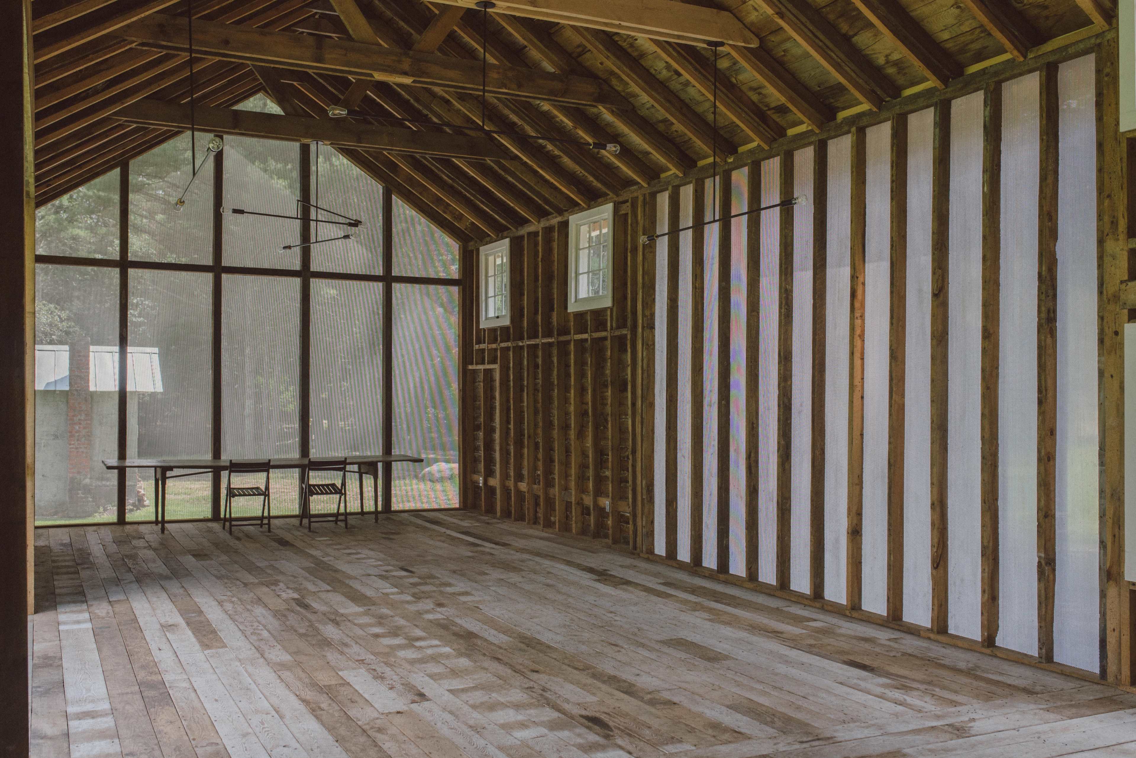 An interior of a spacious, unfinished wooden structure with large windows and a simple table and chairs setup against the wall.