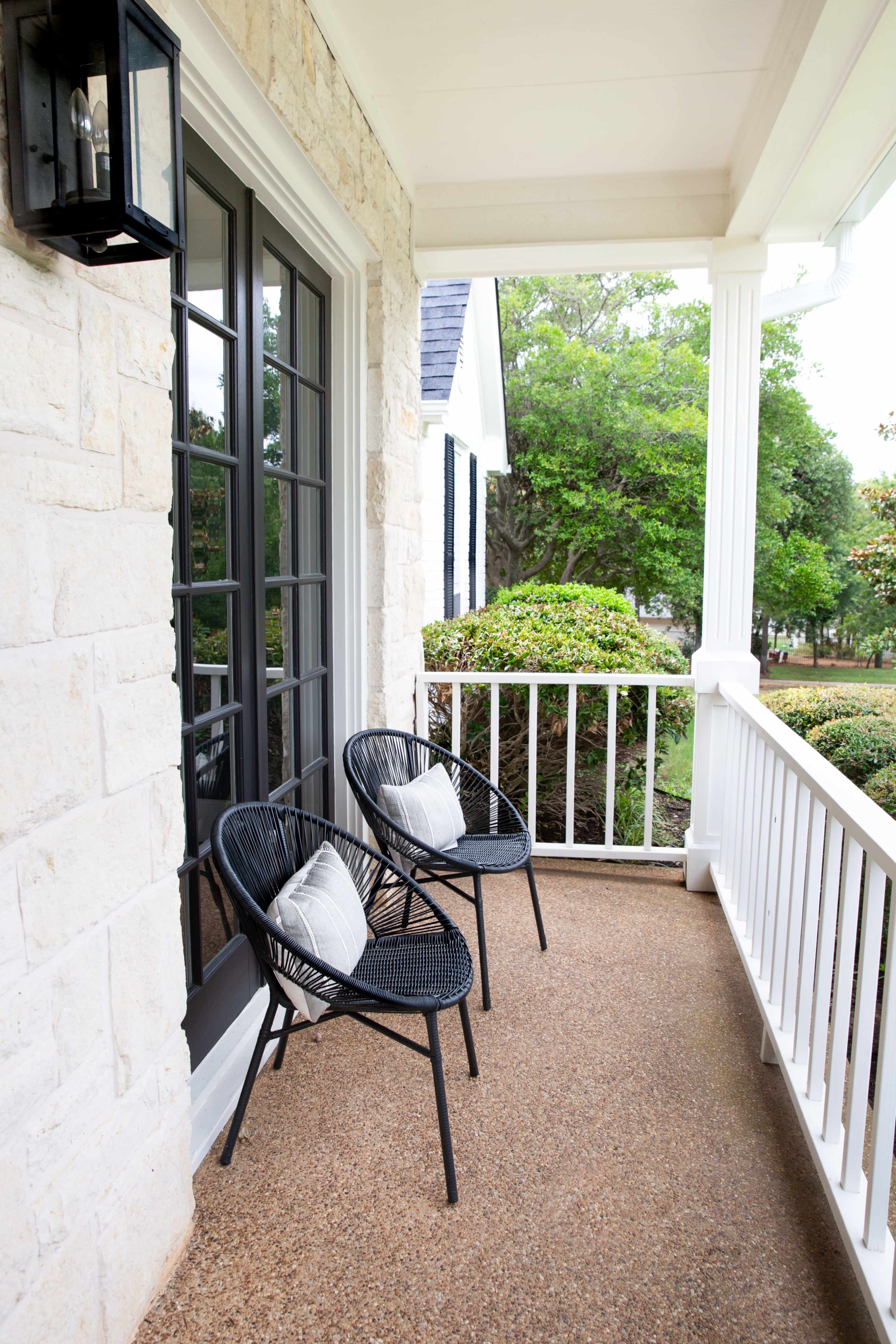 A porch with two black chairs and a small table, surrounded by greenery and a stone wall.