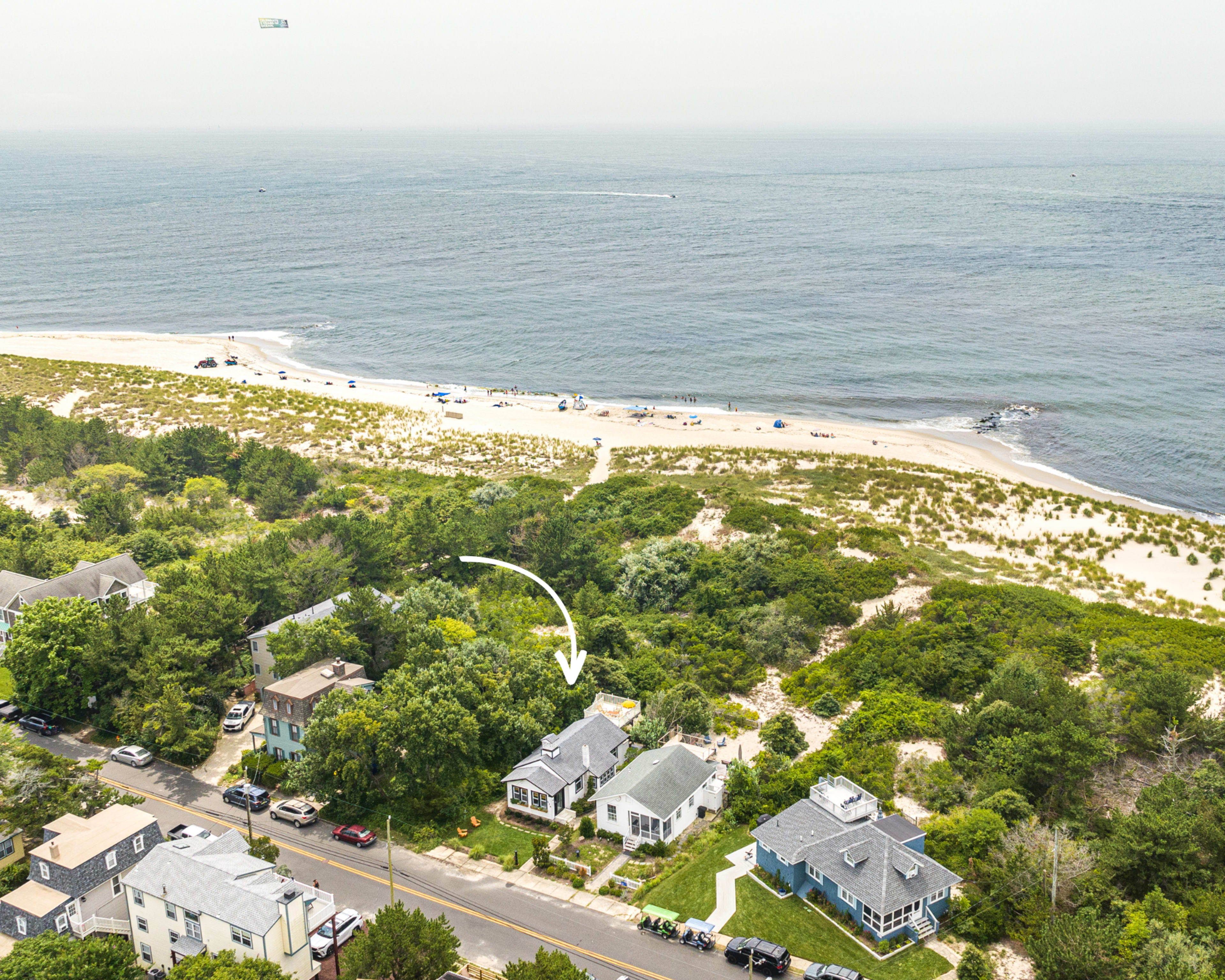 An aerial view shows a beachfront area with a row of houses along a road and a sandy beach in the background.