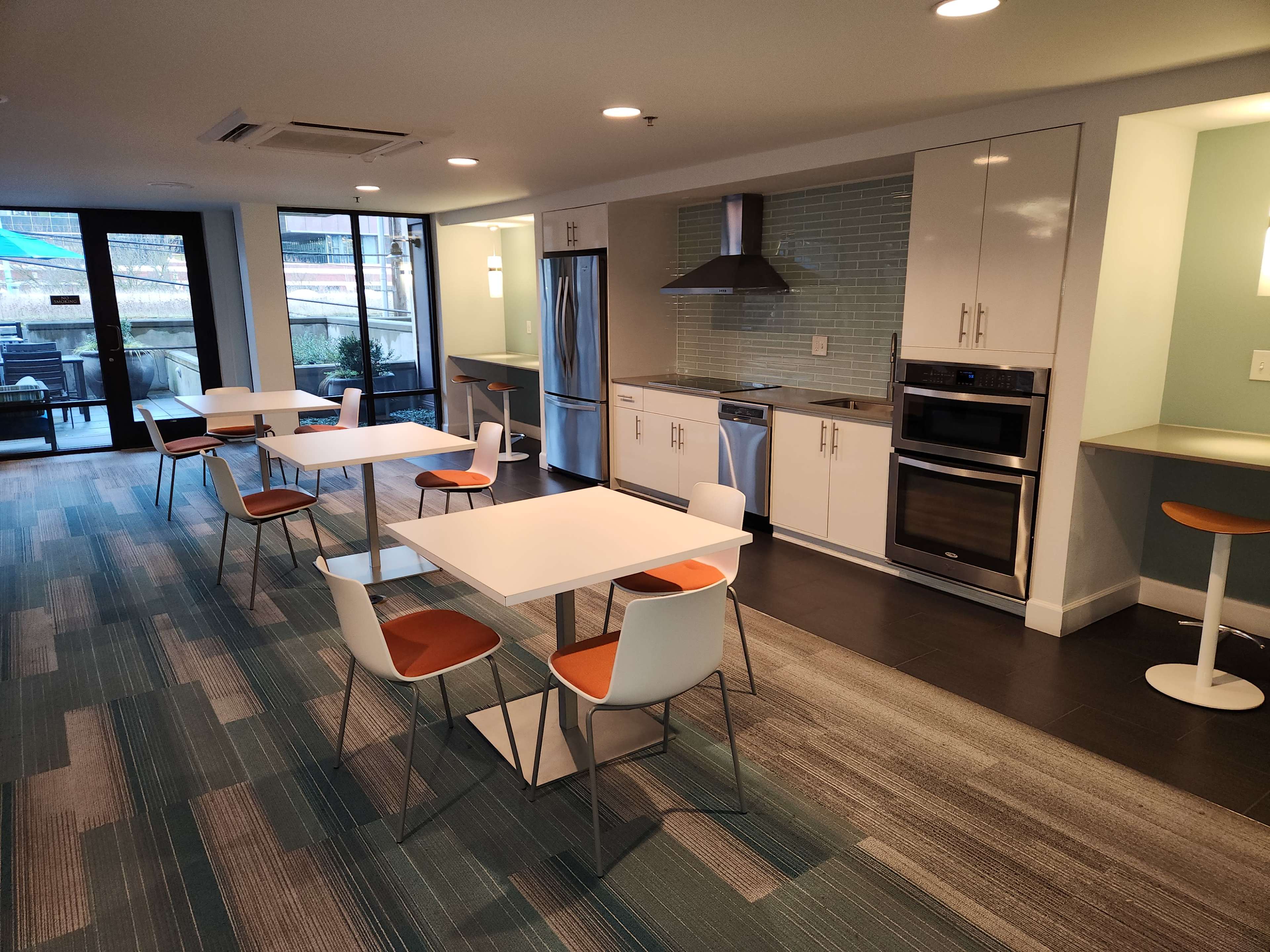 A modern kitchen and dining area featuring stainless steel appliances, white tables with orange-accented chairs, and large windows overlooking an outdoor space.