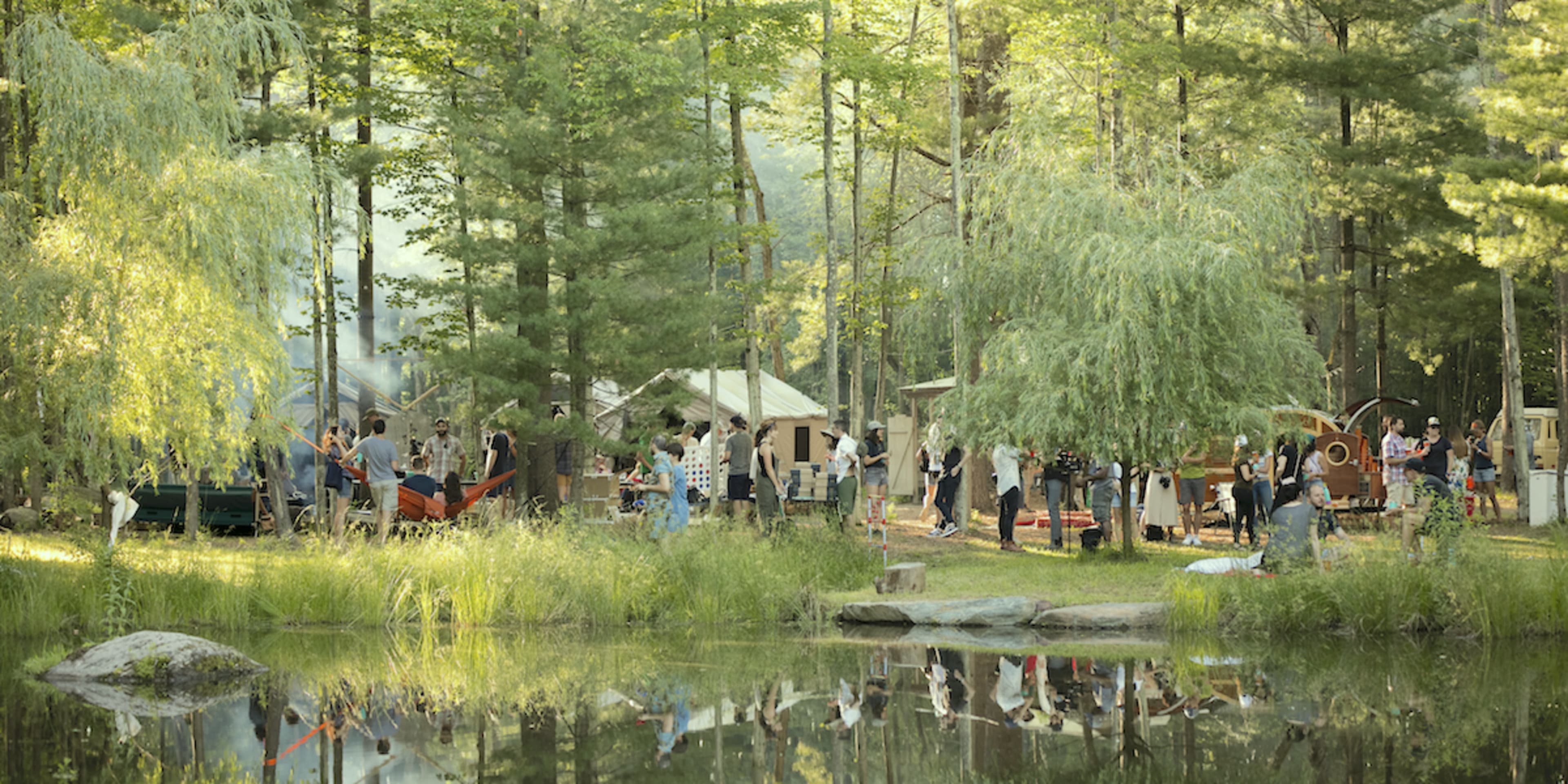 A group of people gathers near a pond surrounded by tents and trees in a forested campsite.