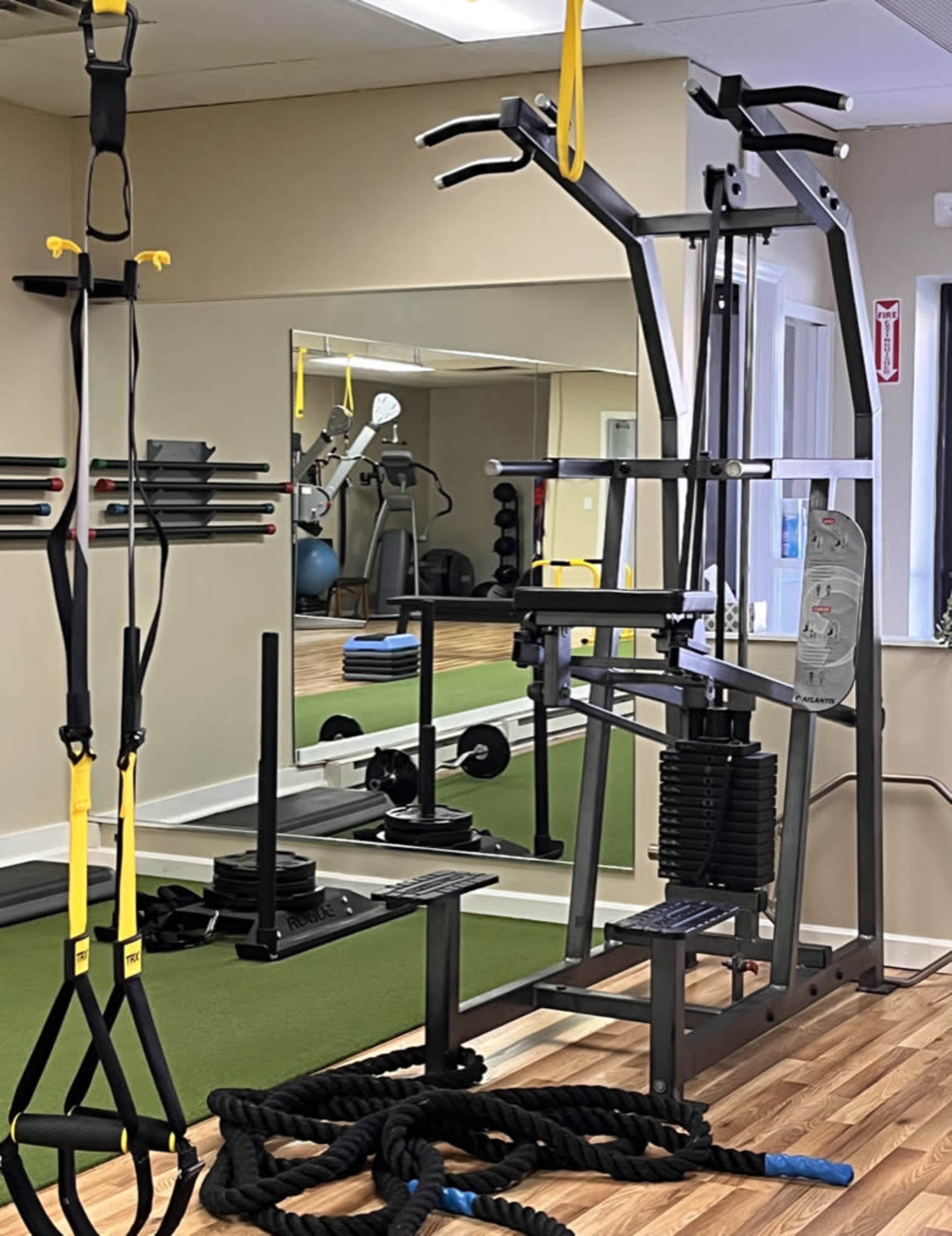 A gym interior featuring exercise equipment, including a multi-station weight machine, resistance bands, and battle ropes, with a mirrored wall reflecting the scene.