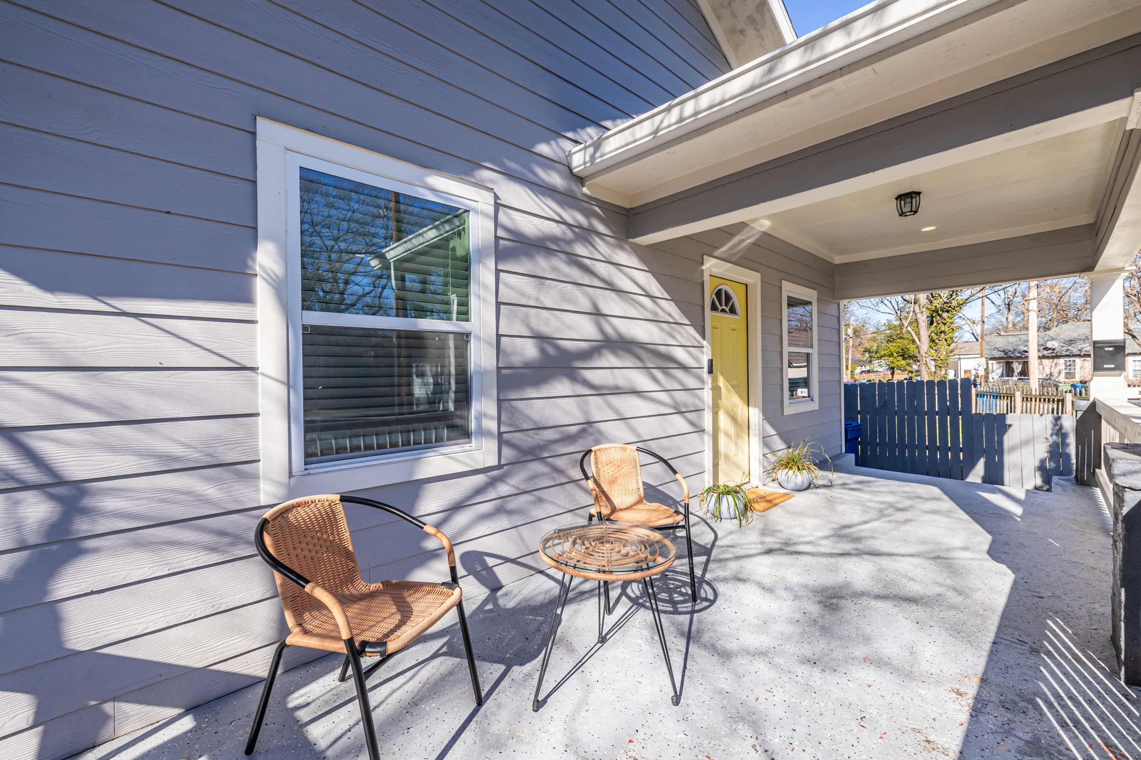 The image shows a porch with two wicker chairs and a small round table, alongside a yellow front door and a gray house exterior.