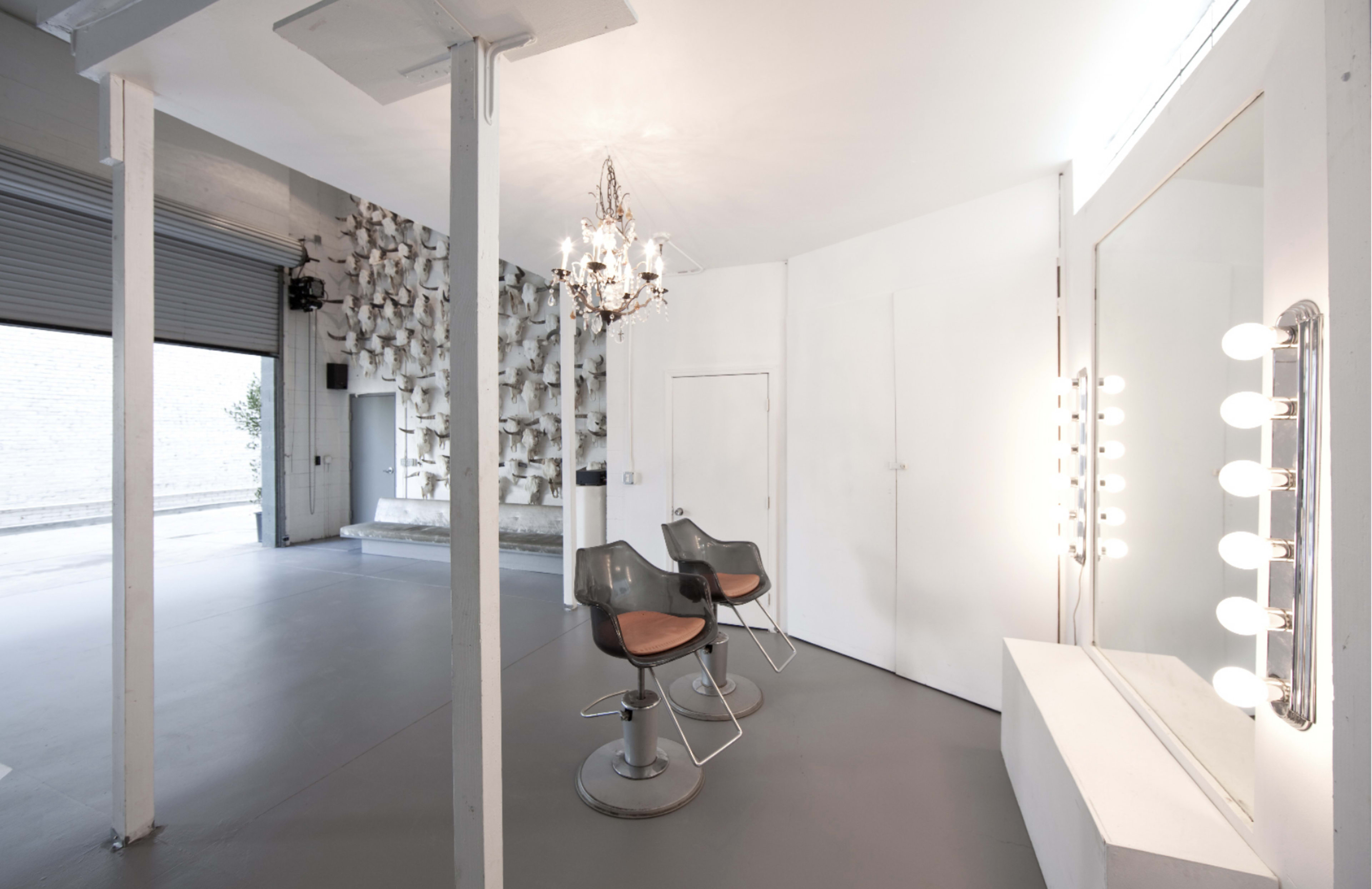 The image shows a modern, minimalistic hair salon with two styling chairs, a large mirror with lights, and a decorative wall feature.