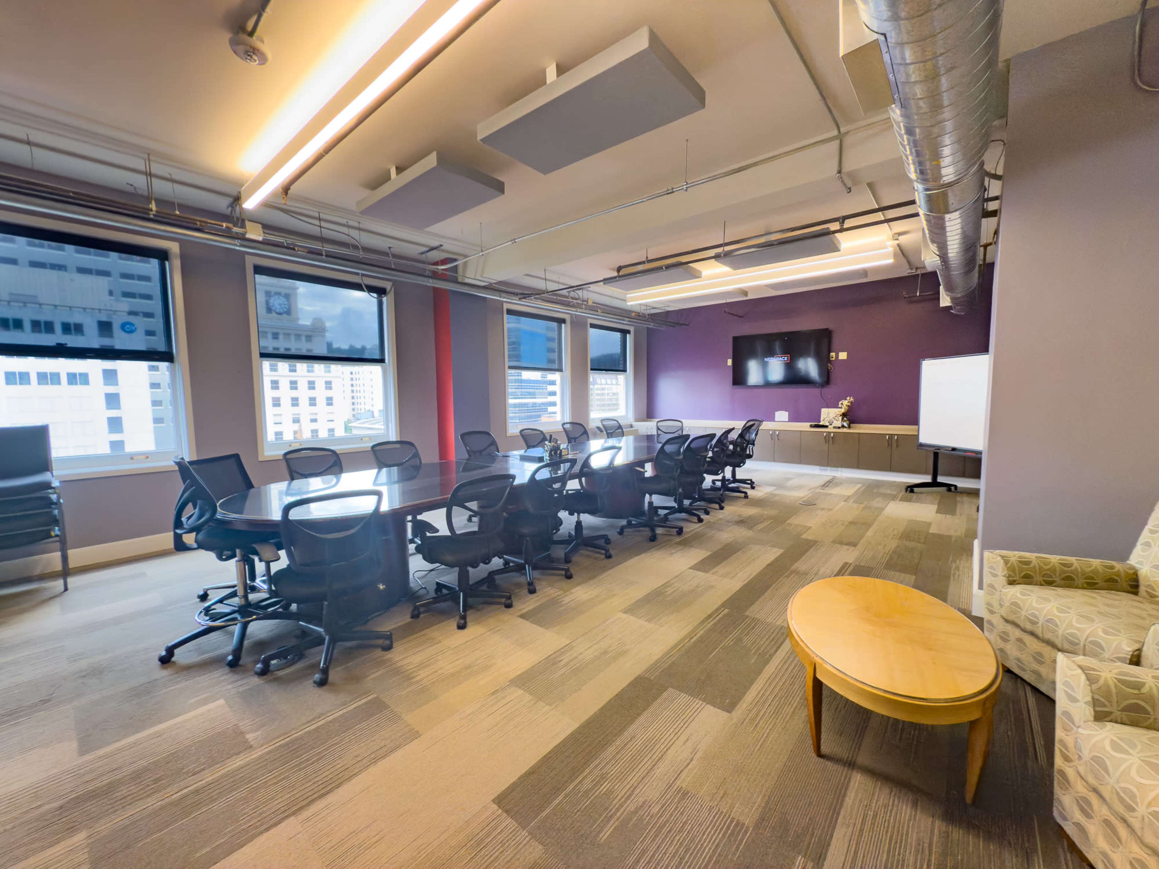 The image shows a modern conference room with a long table surrounded by black office chairs, large windows revealing a city view, and a presentation screen on the wall.